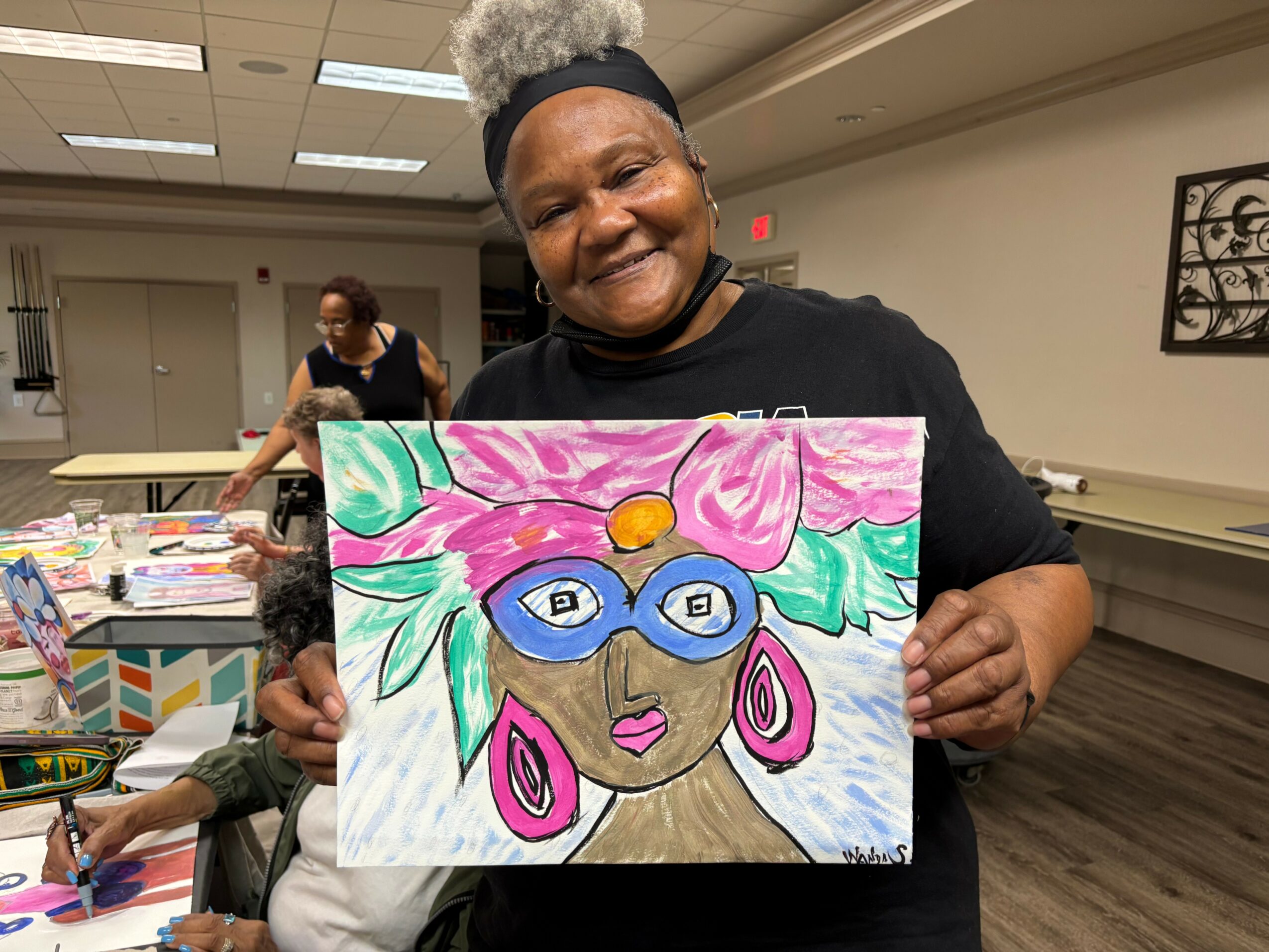 Smiling woman holding colorful painting of a woman with pink hair, blue glasses, and earrings, in a room with other people.