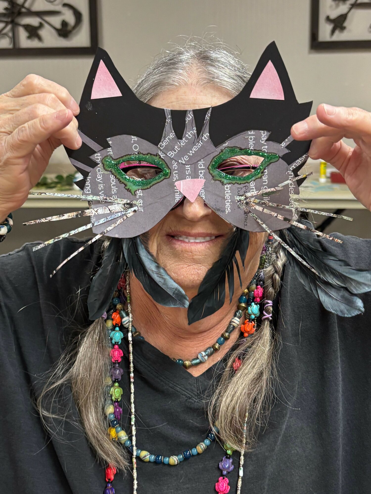 Person holding a black cat-shaped paper craft with colorful decorations, partially covering their face, wearing a black shirt and beaded necklace.