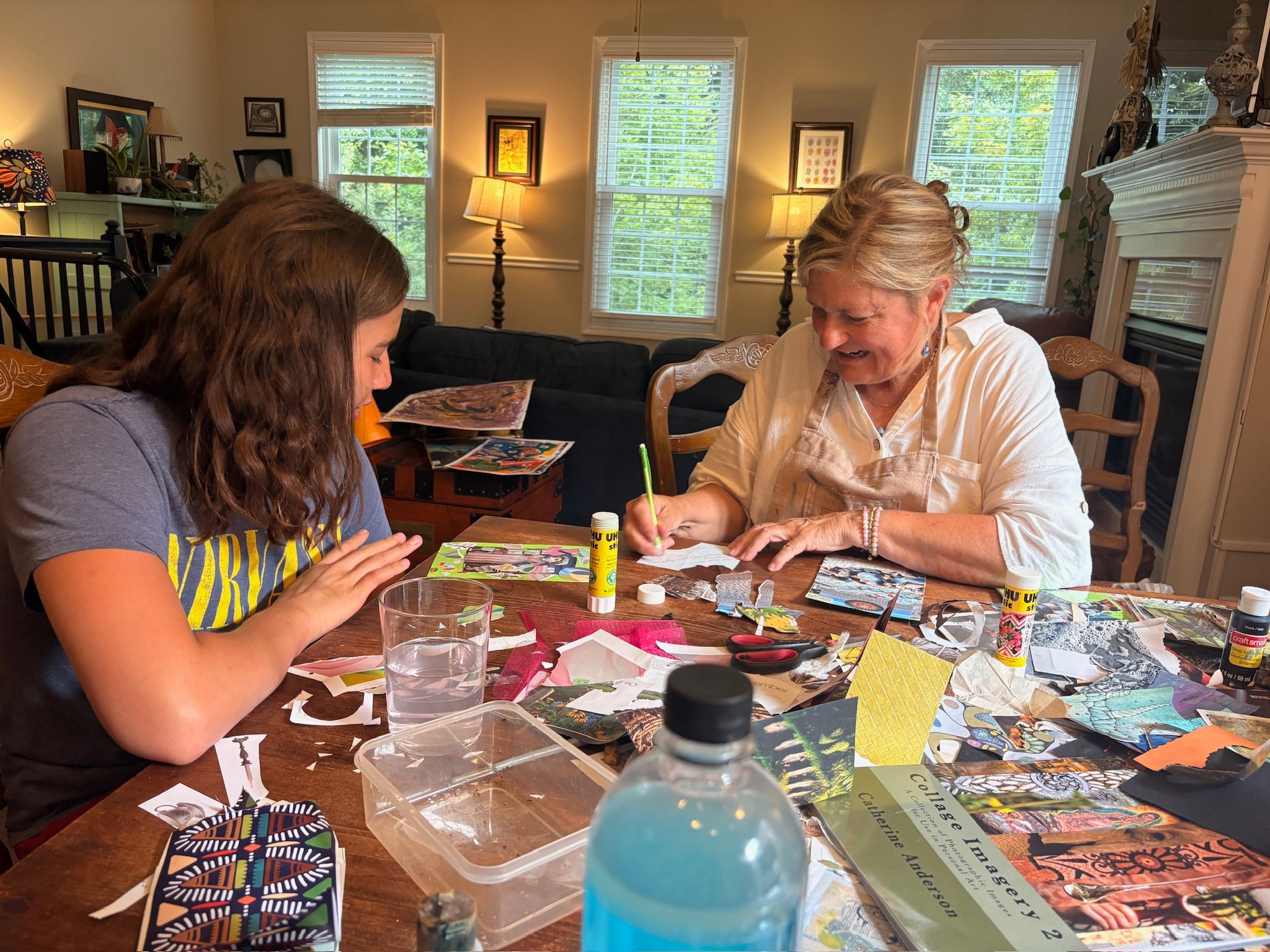 Two women sit at a table engaged in arts and crafts, with various supplies and magazines spread out.