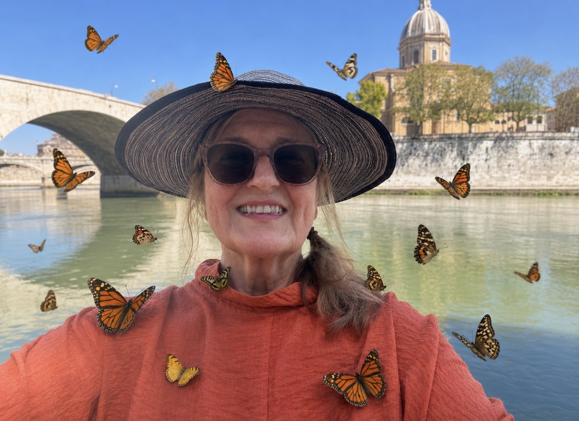 Smiling woman wearing sunglasses and a wide-brimmed hat near a river with a bridge and historic building in background.