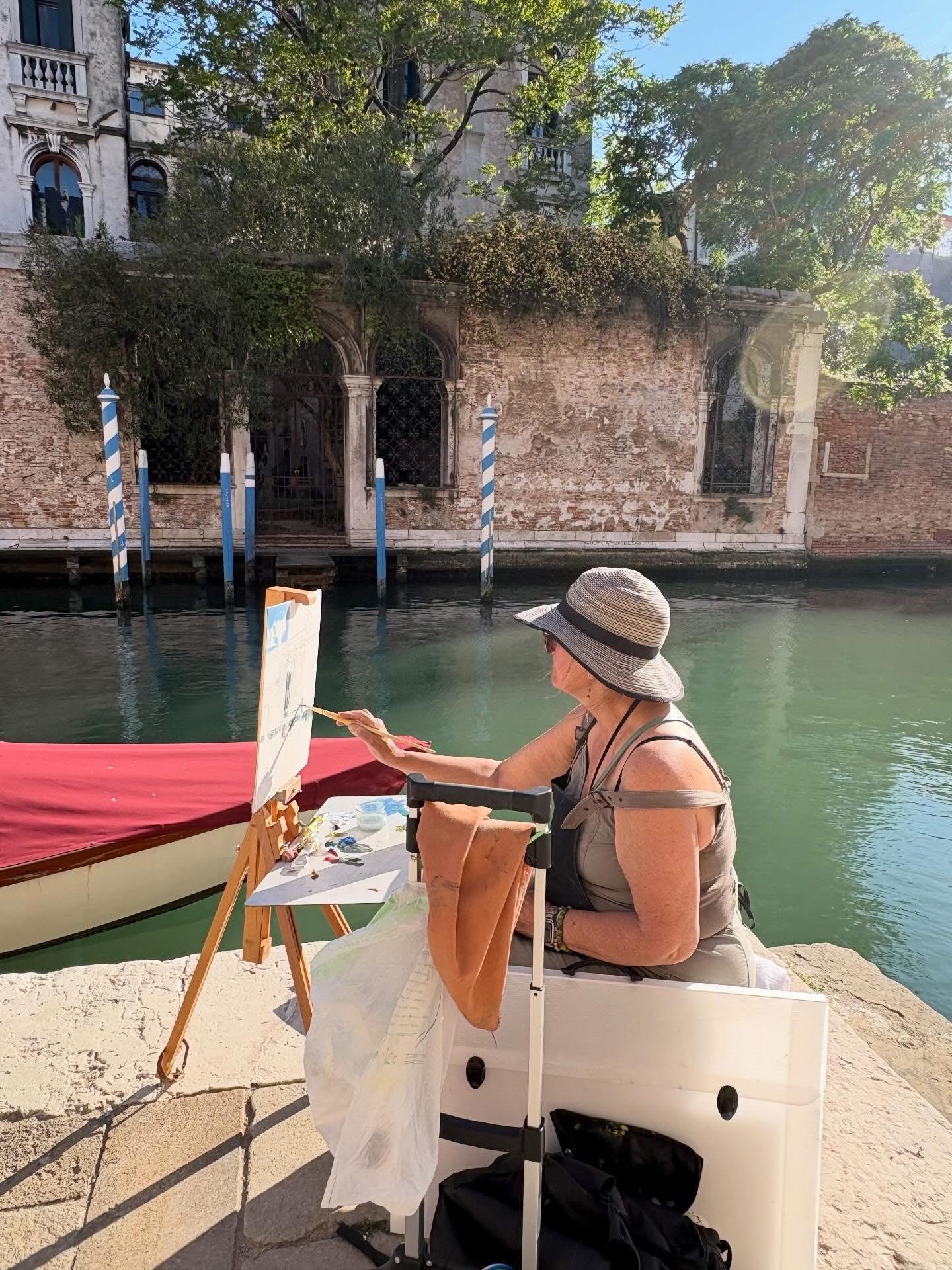 Woman sitting on a white bench by a canal, painting on an easel, with a boat and historic building in background.
