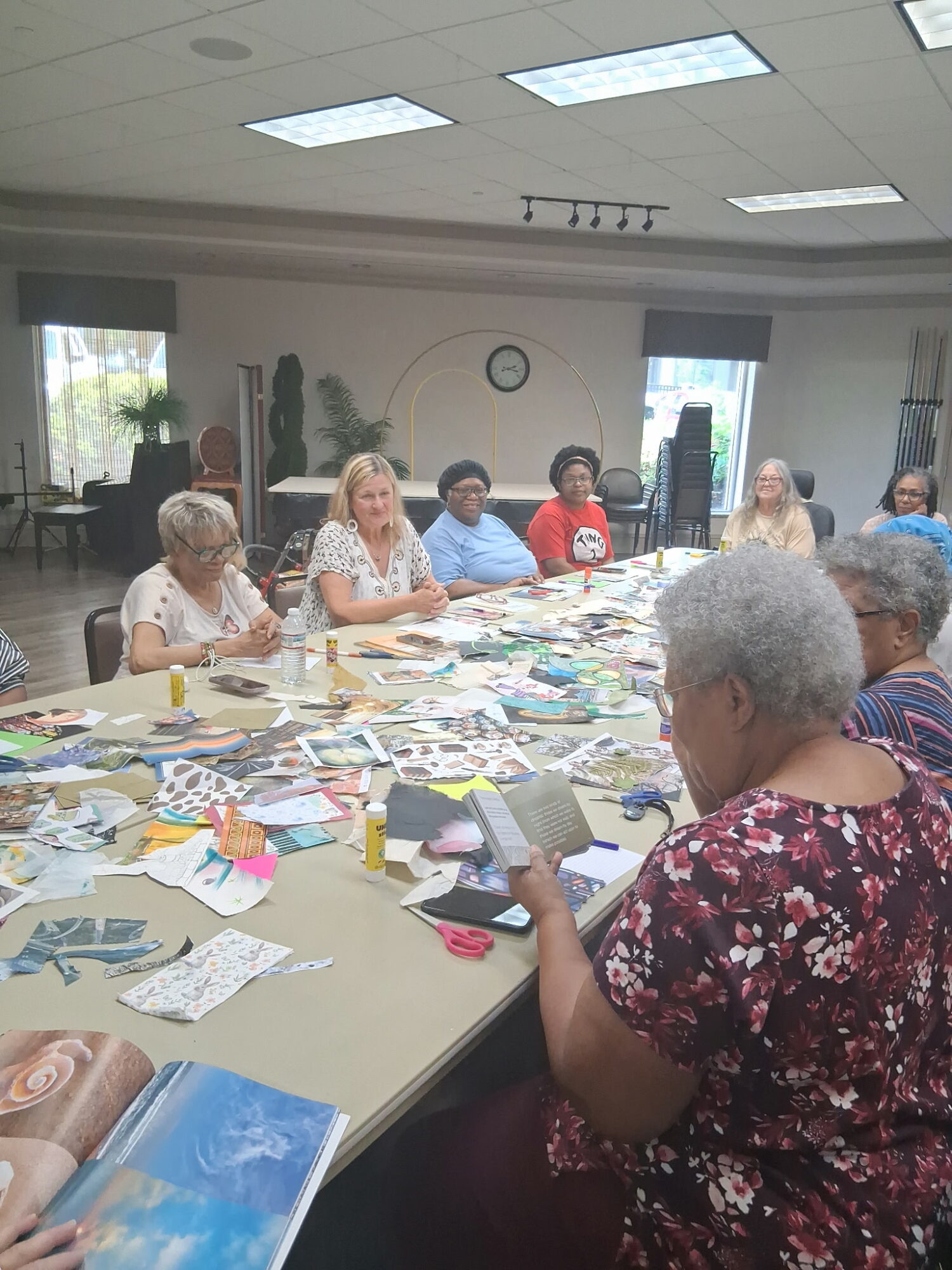 Group of elderly women sitting around a table with craft supplies in a room with windows and ceiling lights.