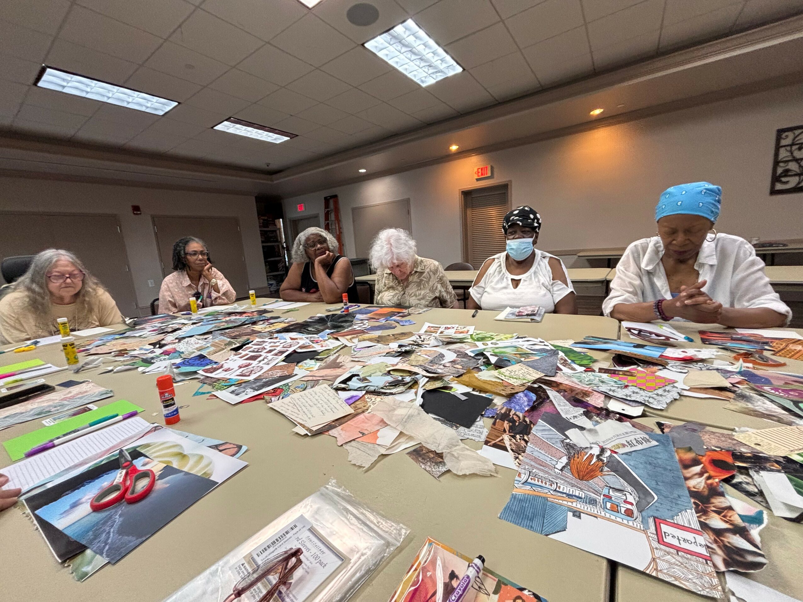 Group of women sitting around a table covered with photos and papers in a room with ceiling lights.