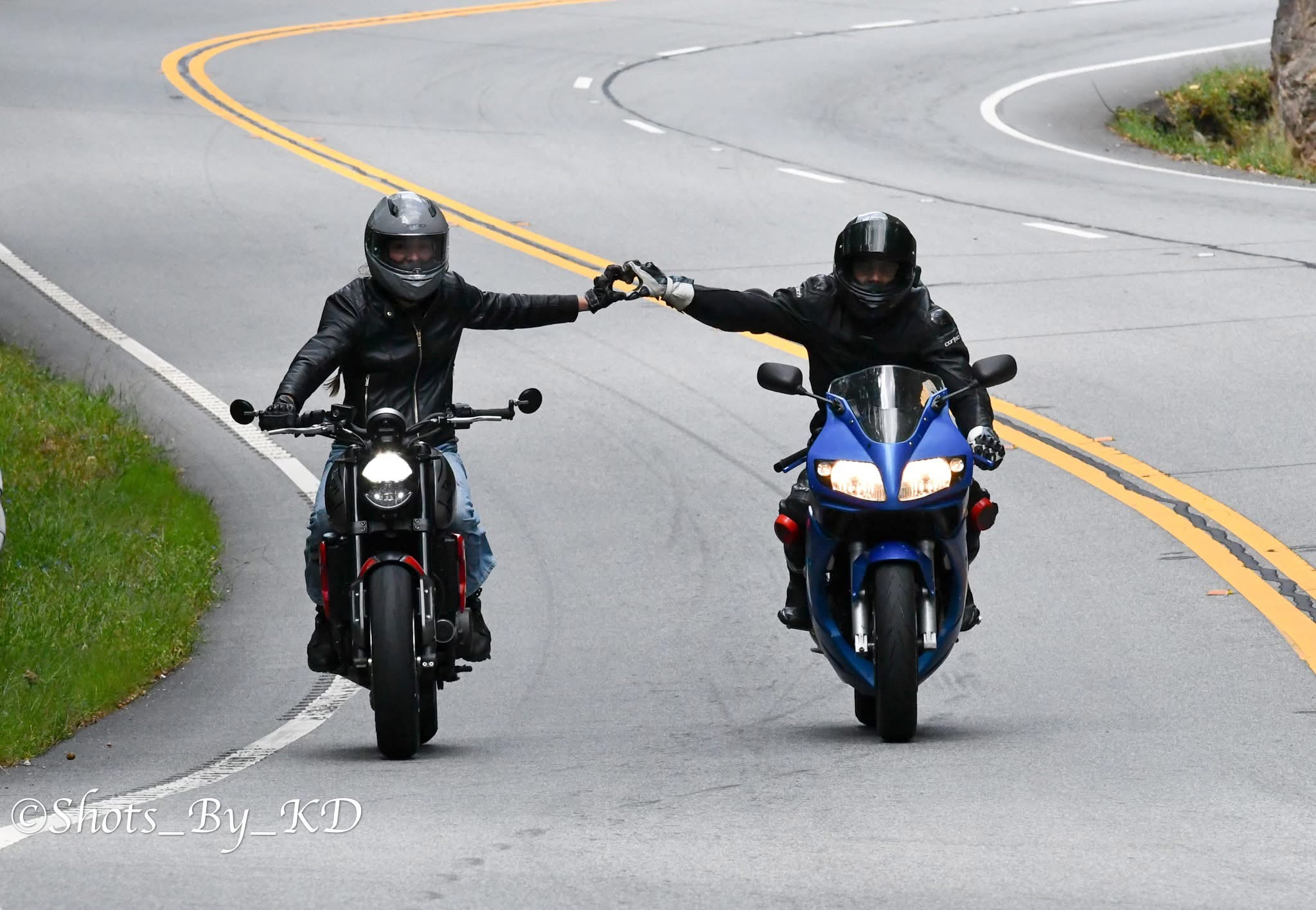 Two motorcyclists wearing black helmets and jackets hold hands while riding on a curved road.