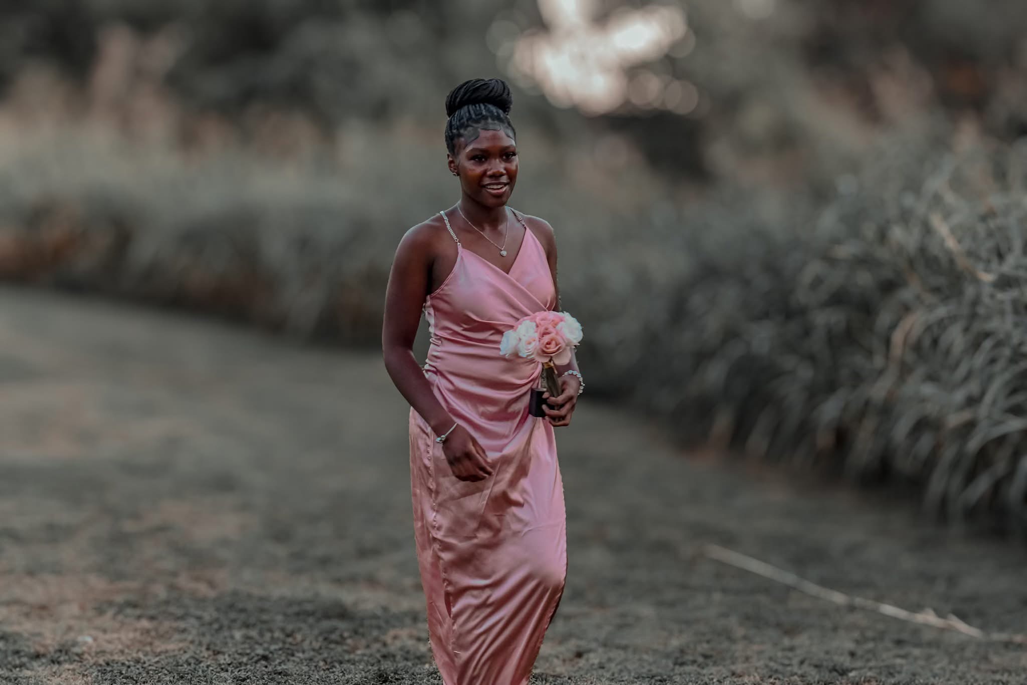 Woman in pink dress holding a bouquet, standing outdoors on a grassy path, smiling, with blurred greenery in background.