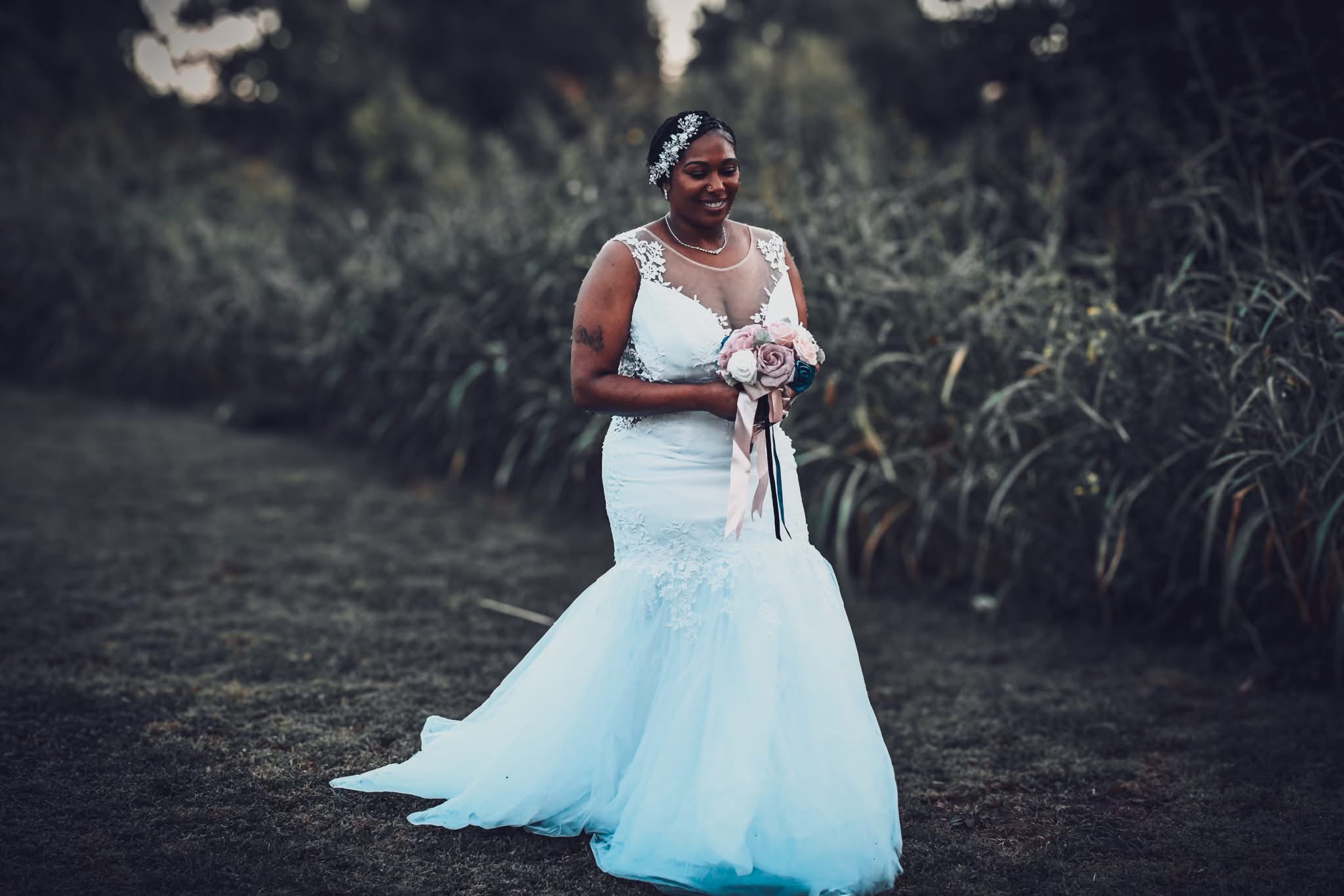 Woman in wedding dress holding bouquet outdoors, smiling, surrounded by greenery