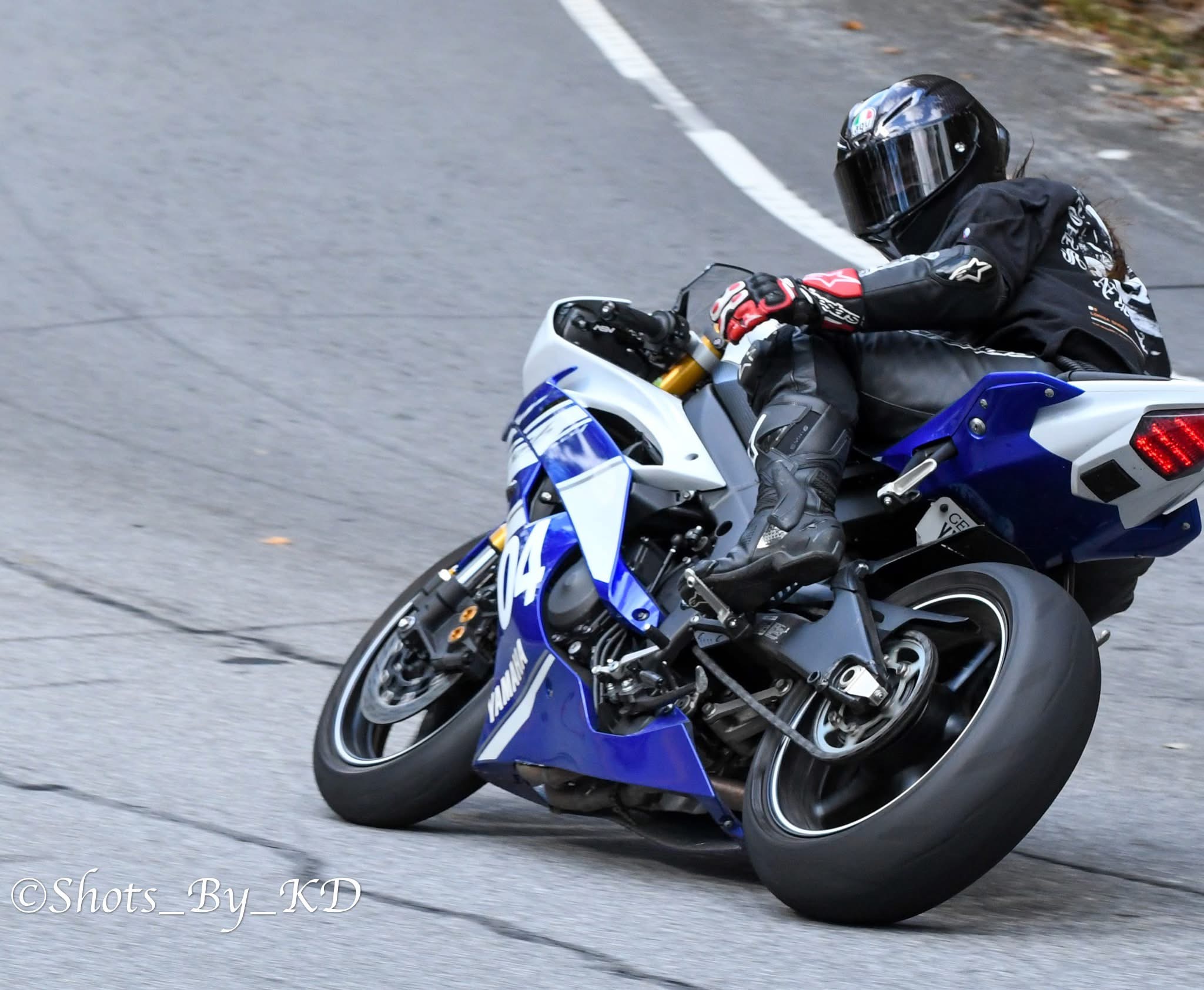 Motorcycle rider leaning into a turn on a curved road, wearing black gear and helmet, blue and white motorcycle.