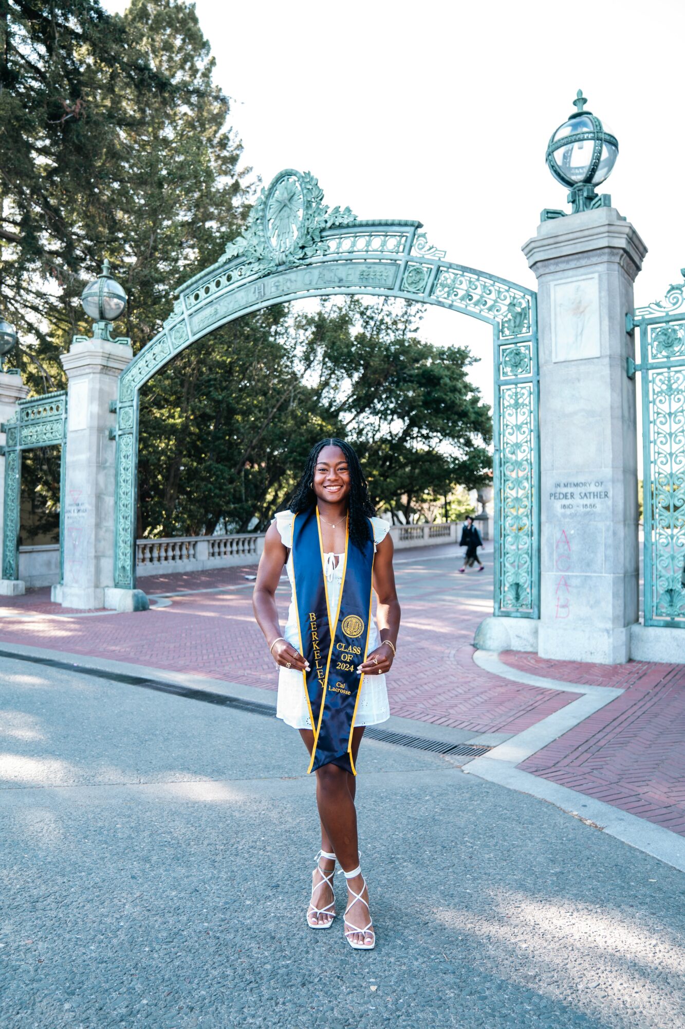 Woman standing outdoors in front of an ornate archway, smiling, wearing a white dress and heels, trees in background.