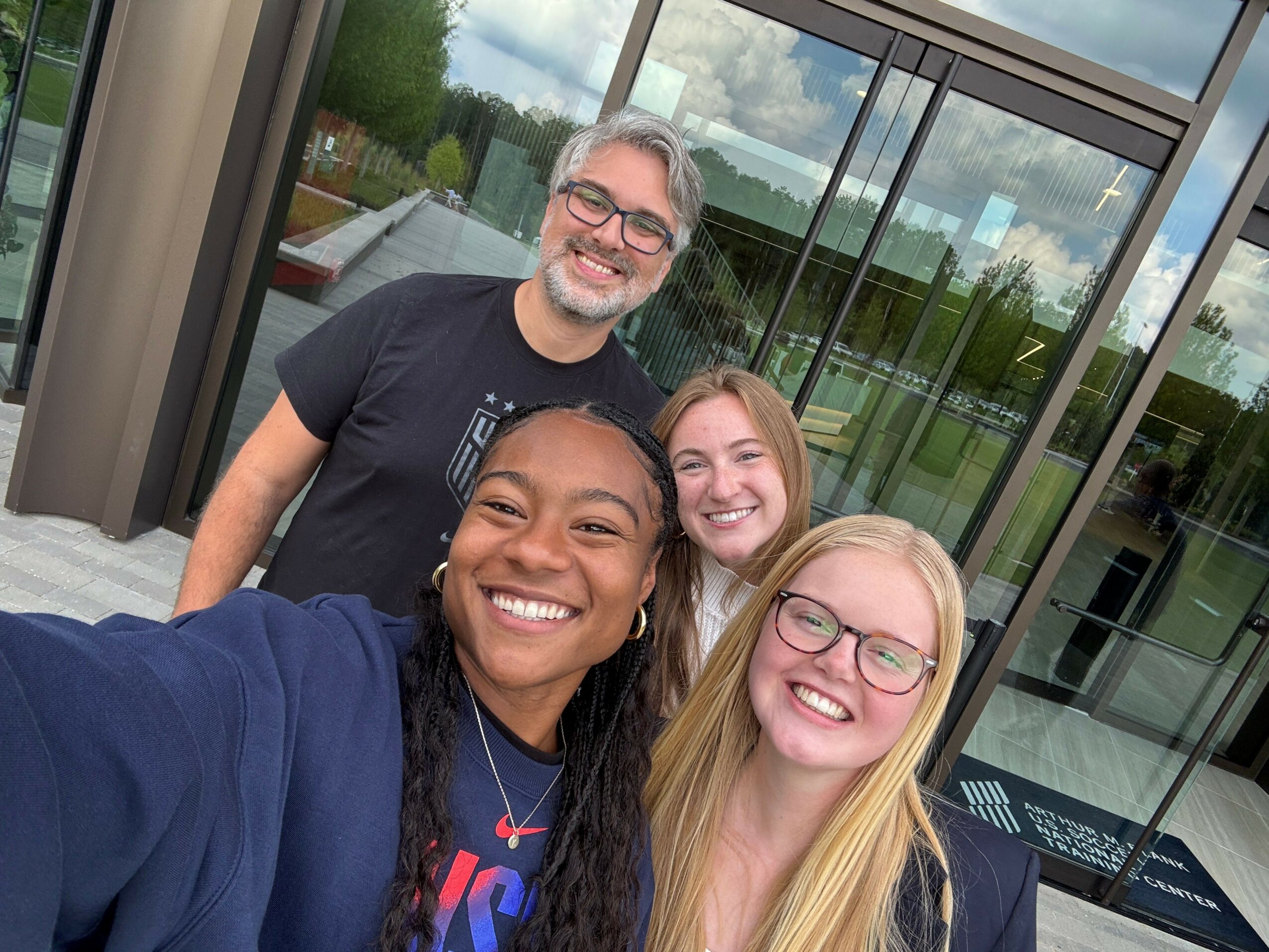 Four people smiling and taking a selfie outside a building with glass windows.