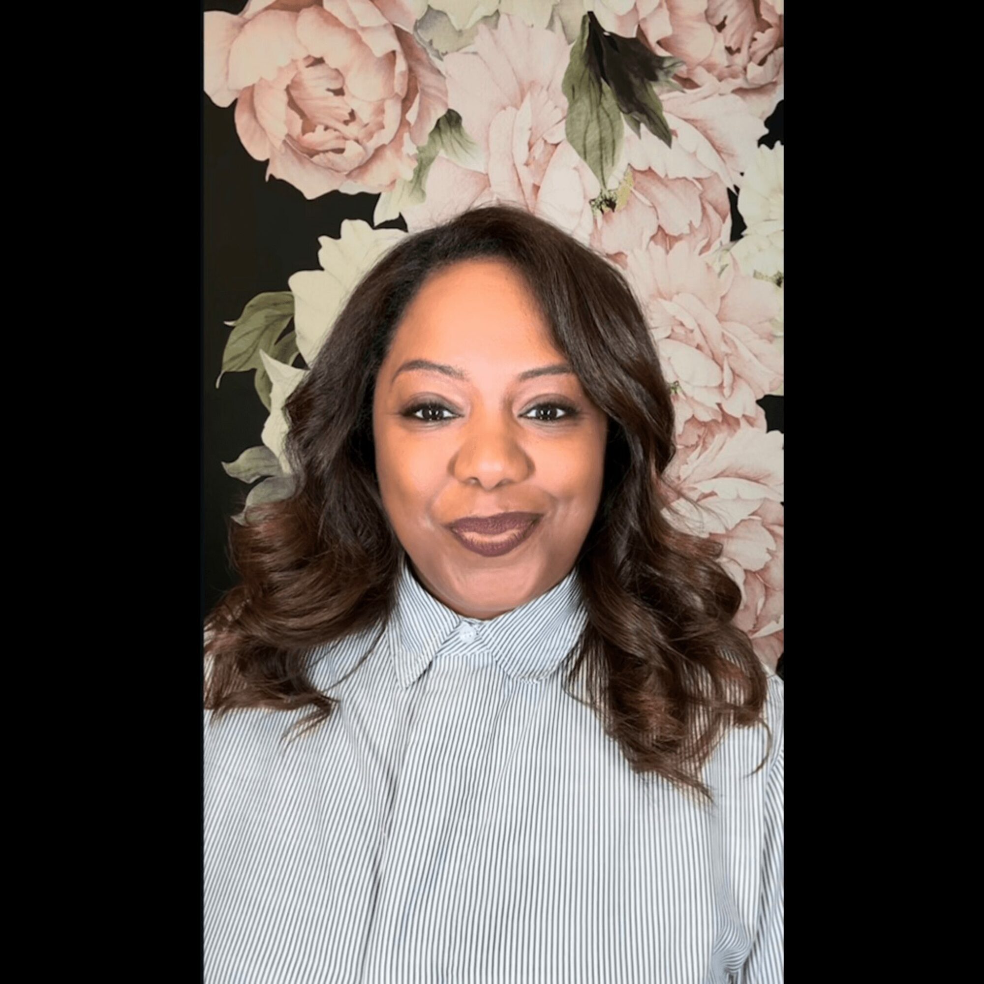 Woman with wavy hair and makeup, wearing a light-colored striped shirt, standing in front of a floral background.