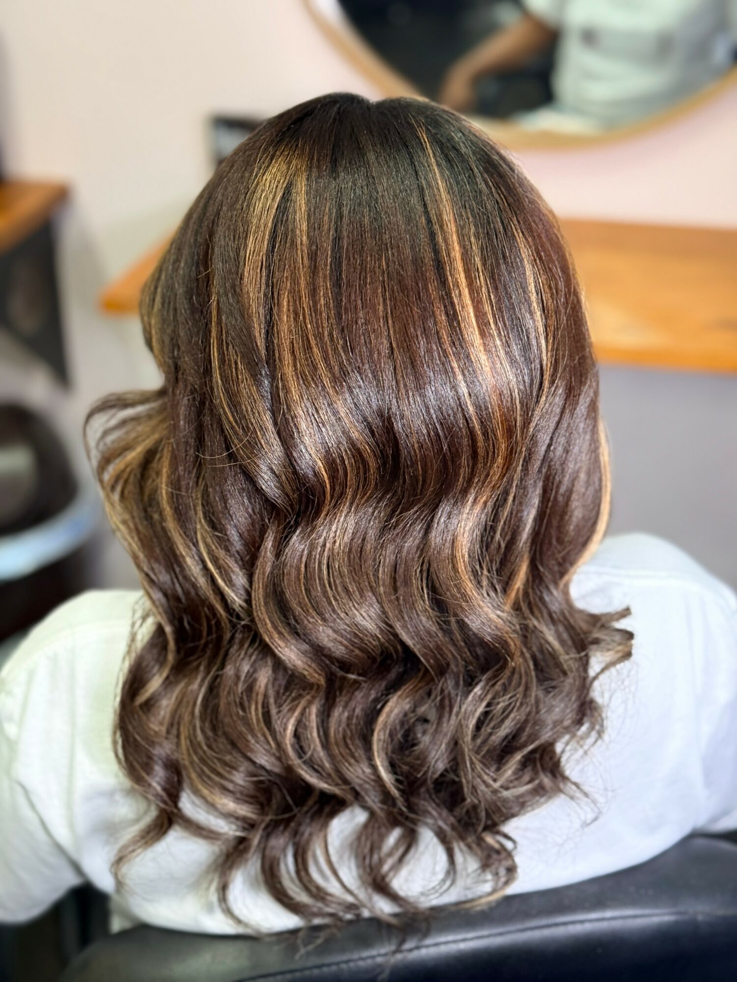 Back of a person's head with wavy, brown hair with highlights, sitting in a salon chair, mirror in background.