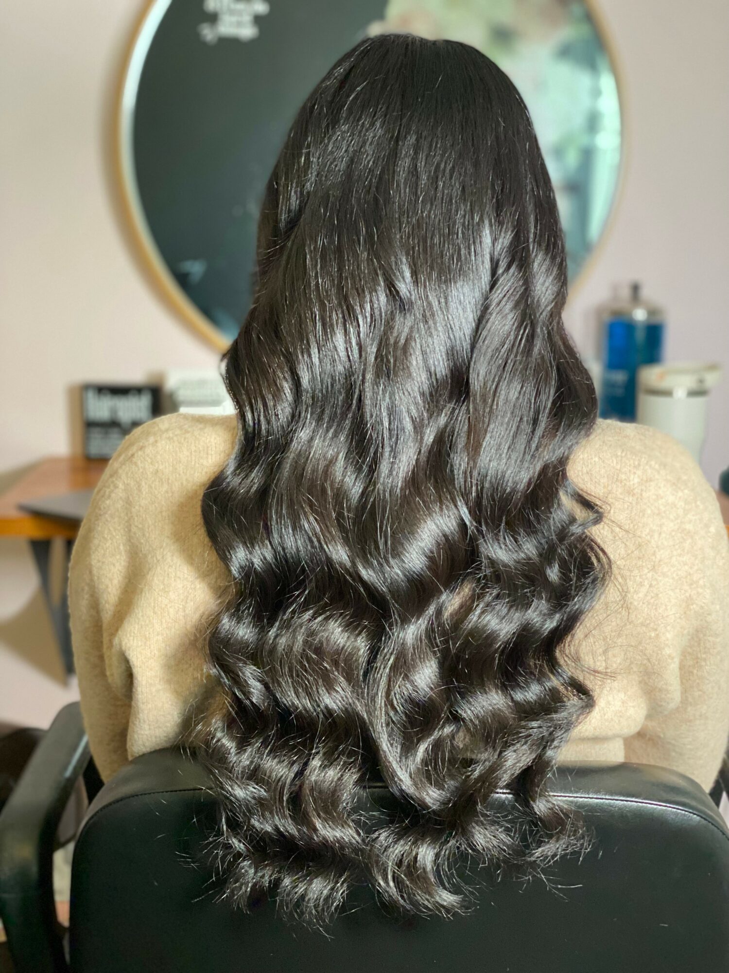 Back of a person with long, wavy, dark hair sitting in front of a mirror in a room.