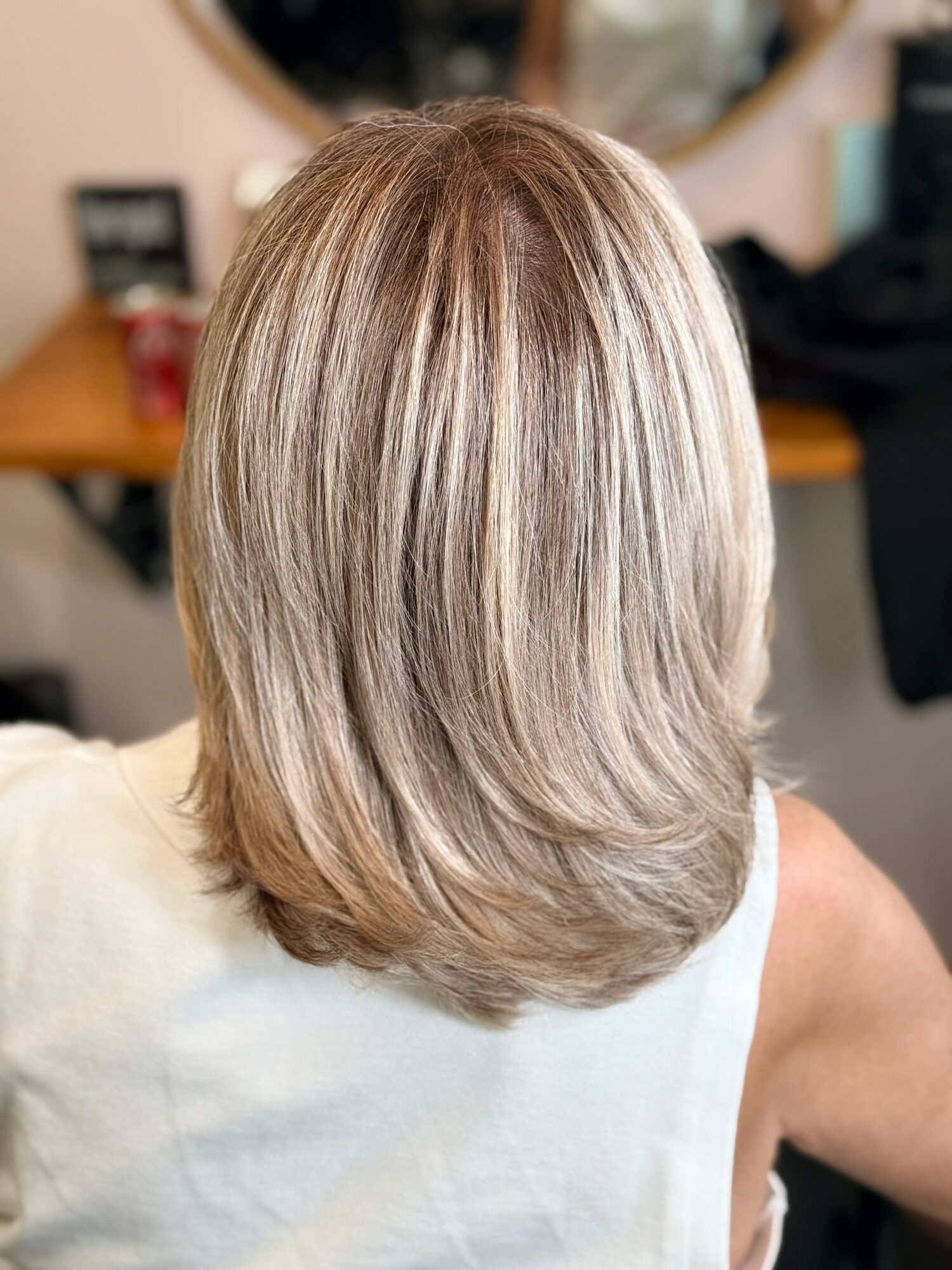 Back view of a person with shoulder-length blonde hair, layered and styled straight, in a salon setting.