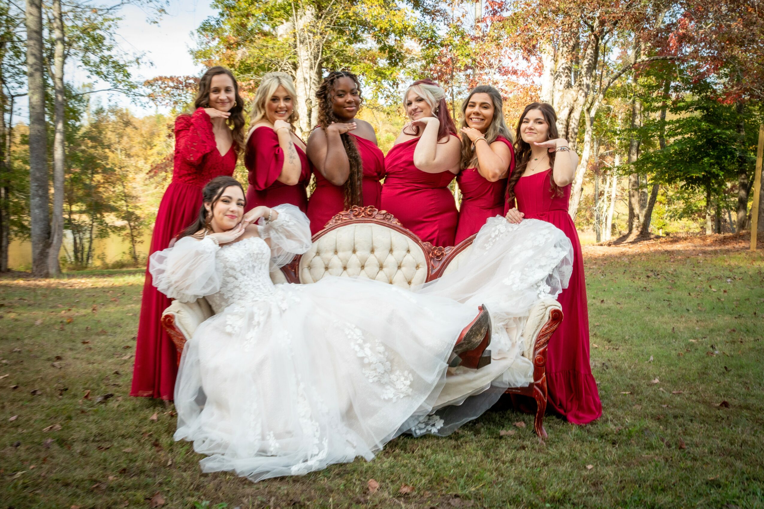 Group of women in red dresses and a woman in a white dress sitting on a bench outdoors, surrounded by trees.
