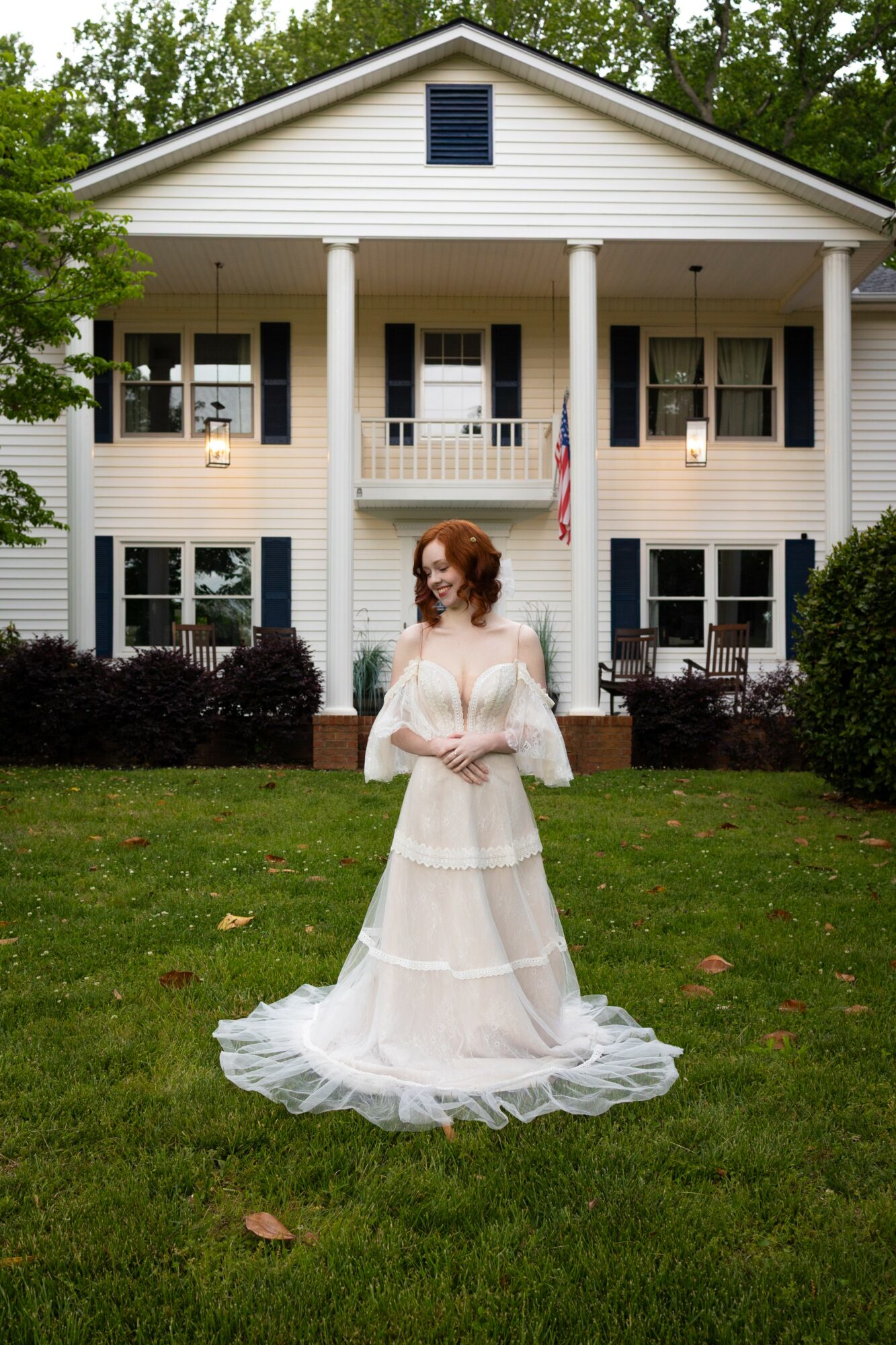 Woman in a white dress stands on grass in front of a large white house with columns and an American flag.