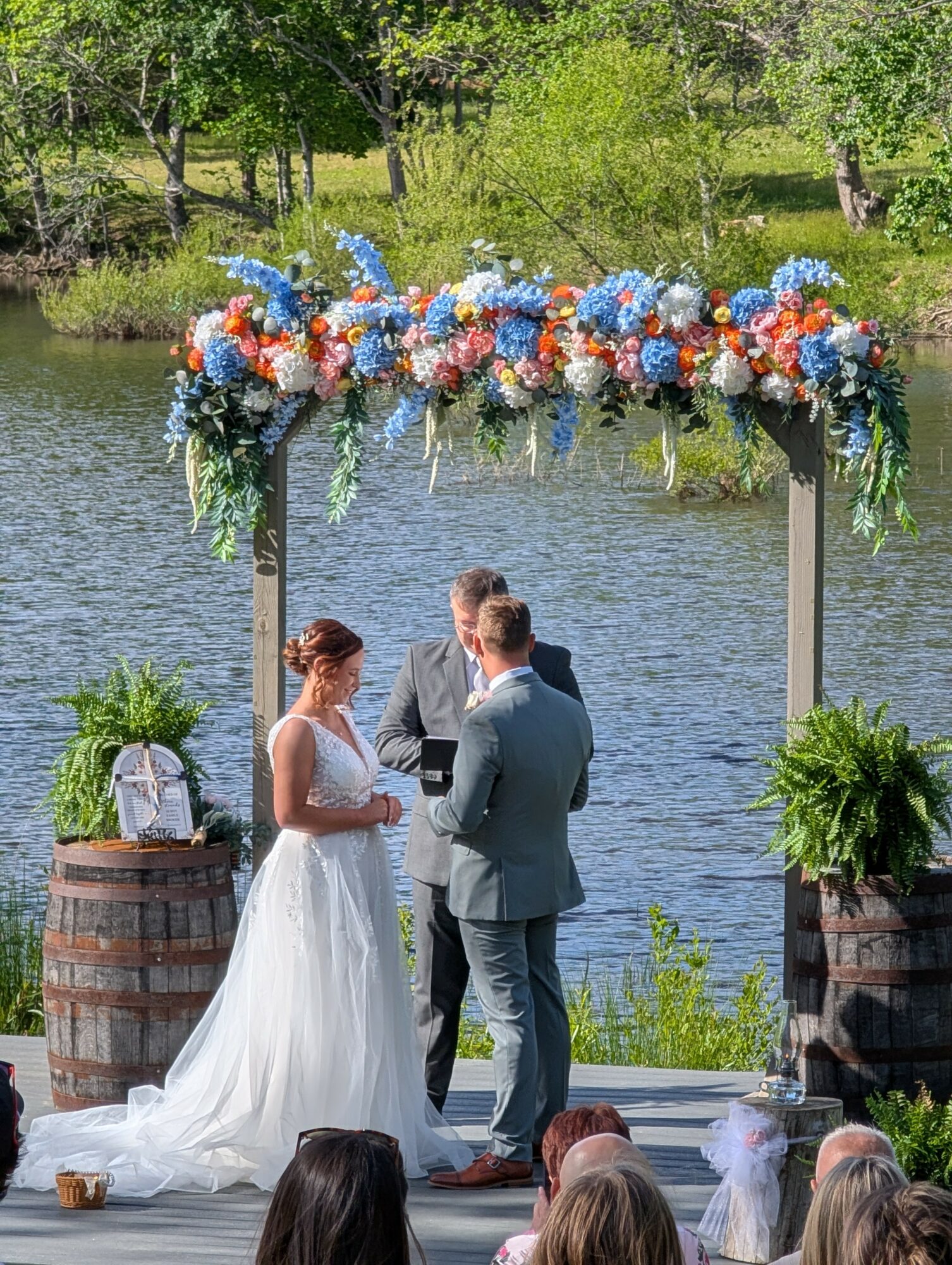 A wedding ceremony with a couple standing under a floral arch by a lake, officiant present, guests seated nearby, trees in background.