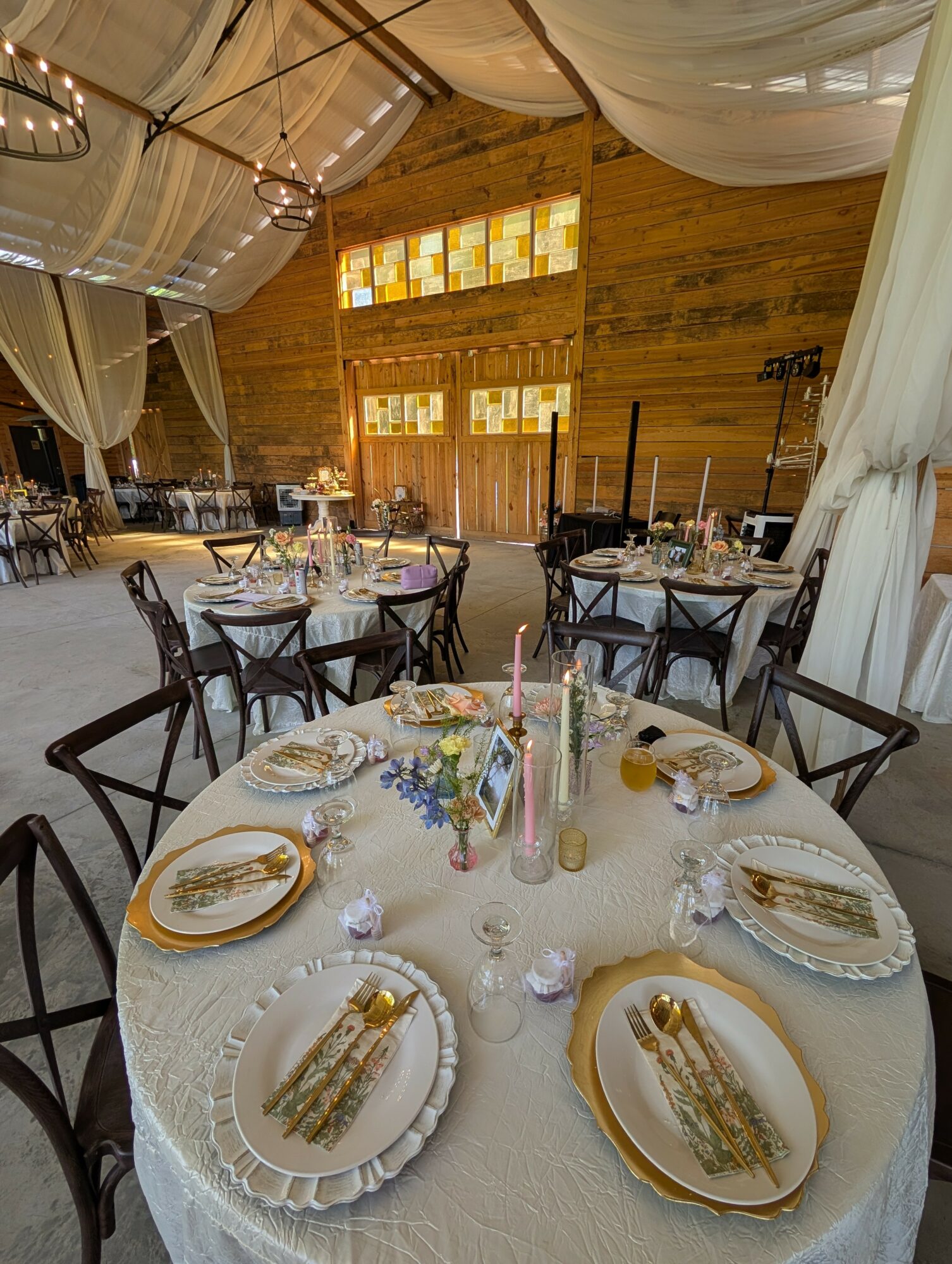 Round table set for a celebration with plates, cutlery, candles, and floral decorations in a rustic venue.