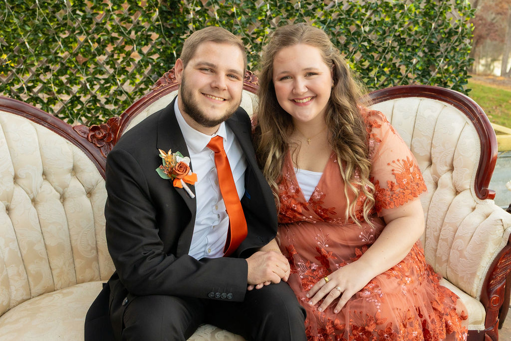 A smiling couple sitting on a vintage sofa outdoors, dressed formally, with greenery in the background.