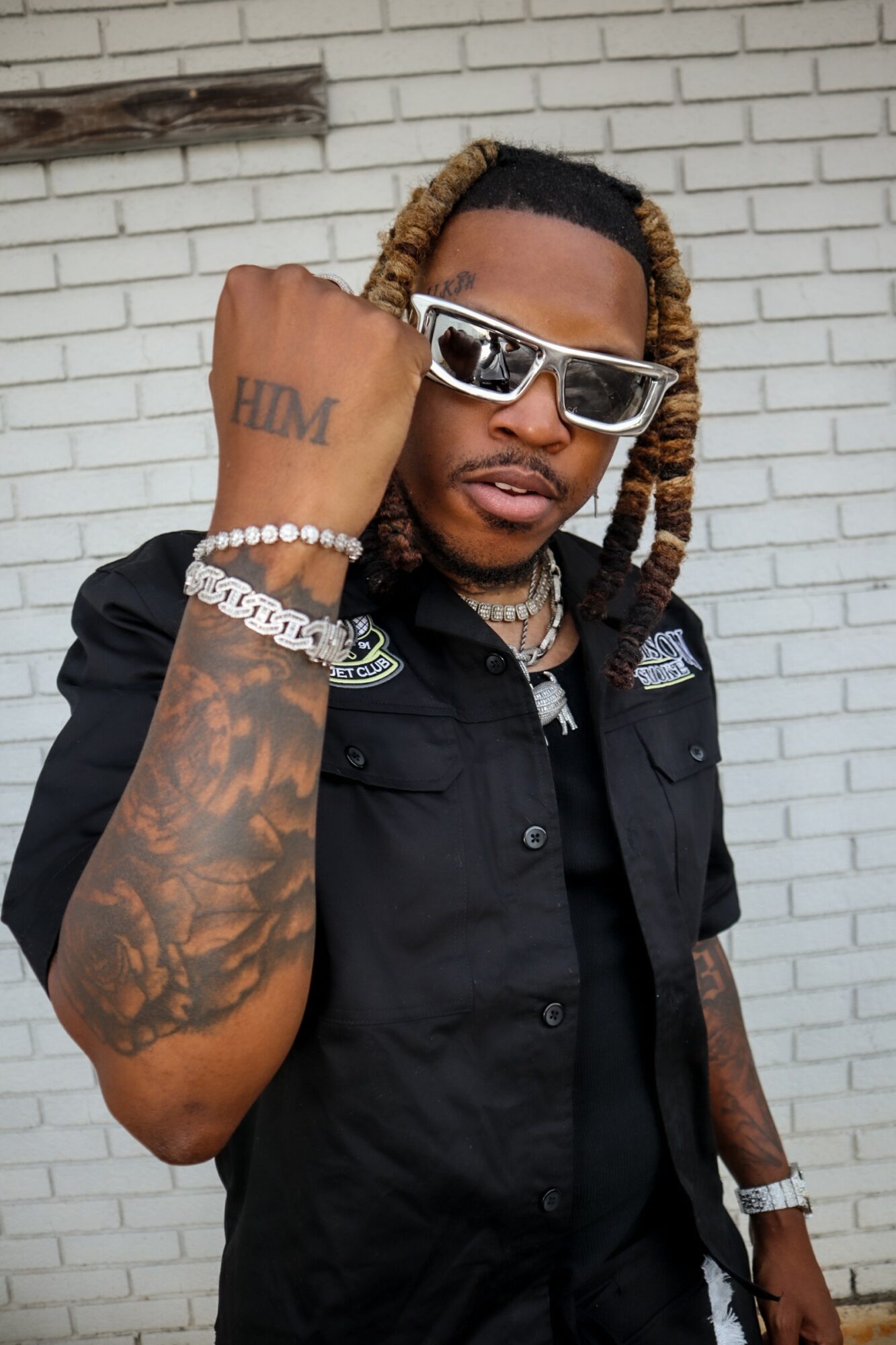 Person with sunglasses and jewelry, showing a tattooed arm, standing against a white brick wall.