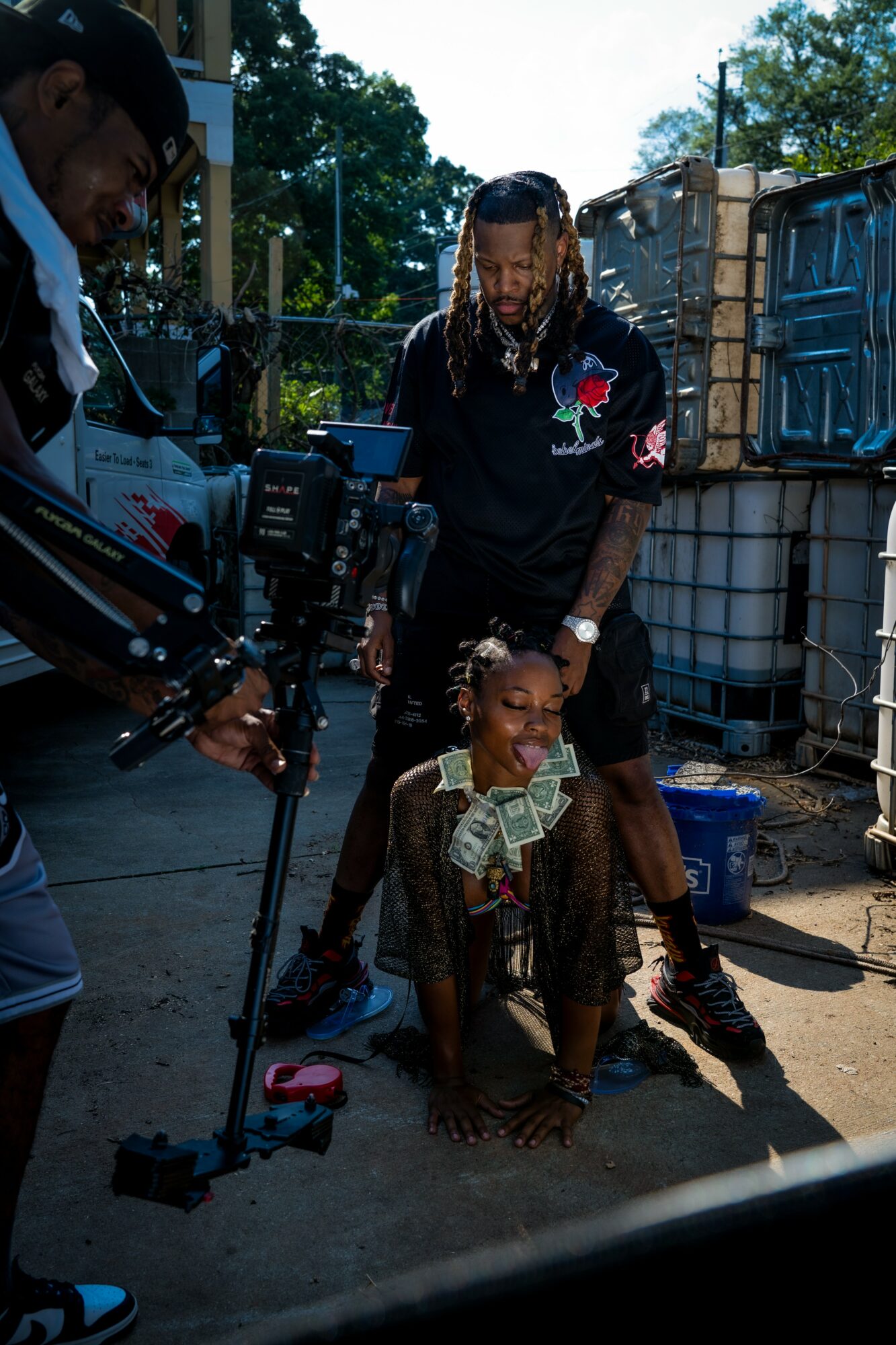 A woman kneels on the ground with money in her hand, smiling, while a man stands behind her, and a person films them.