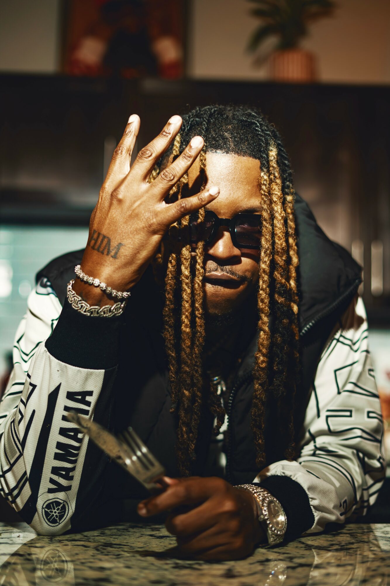 Man with long dreadlocks wearing sunglasses and a patterned jacket, resting his hand on his face, in an indoor setting.