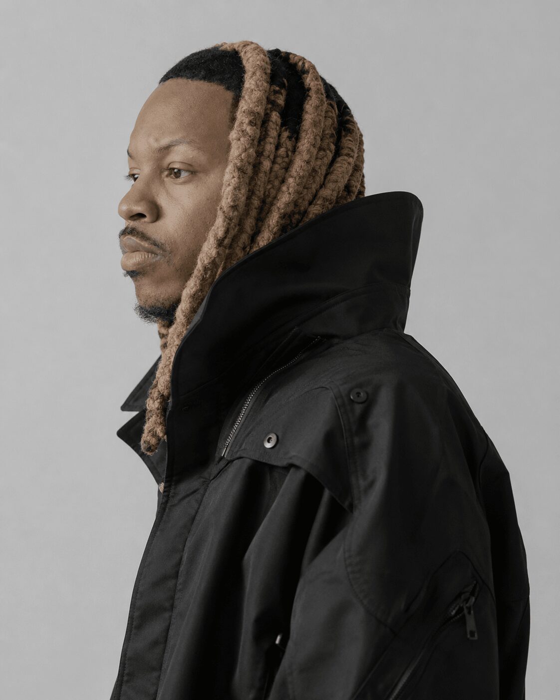 Man with dreadlocks wearing a black jacket, facing left, against a plain background.