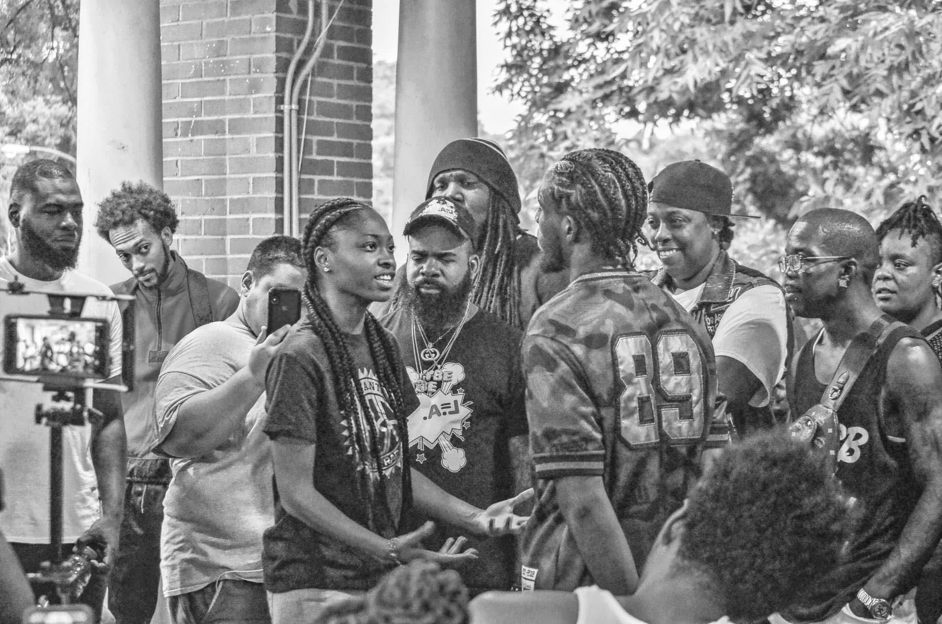 Group of people outdoors, some talking and others listening, with trees and a brick building in the background.