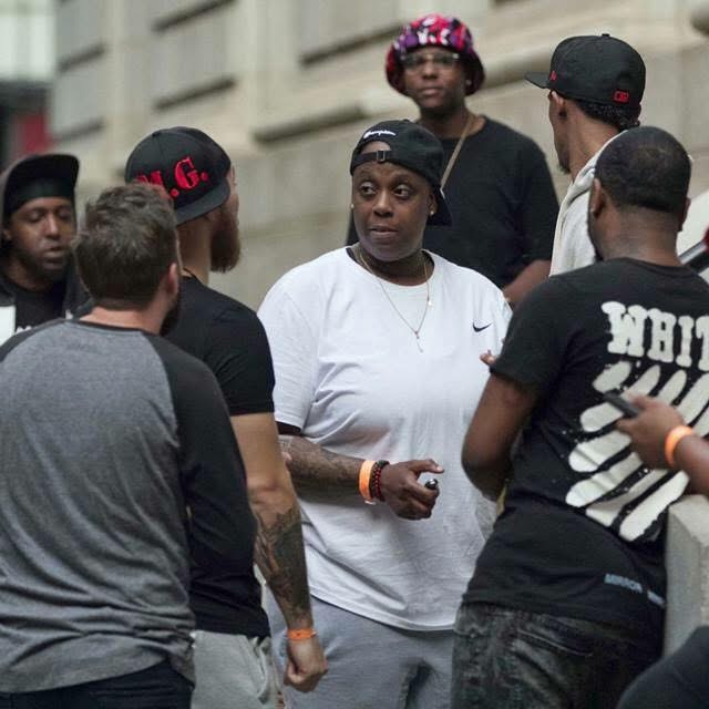 Group of people gathered outdoors, some facing each other, with a woman in a white shirt at the center, wearing a black cap. Word on shirt partially visible.