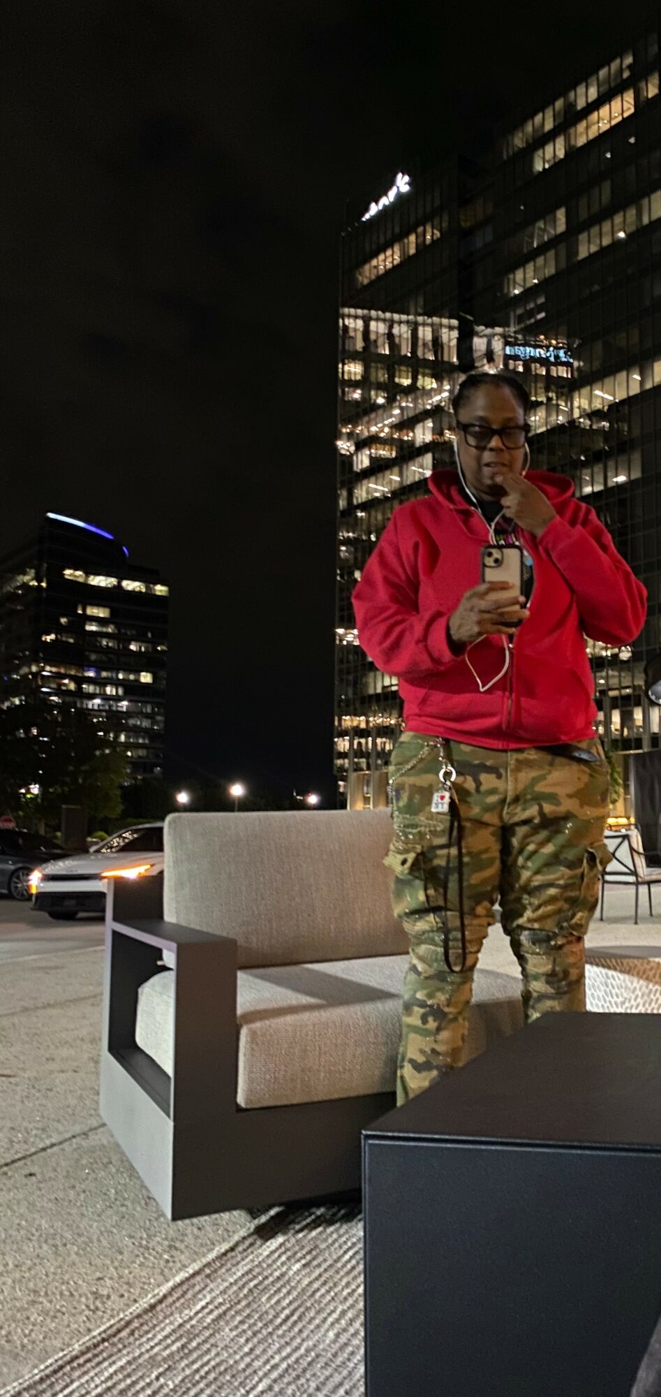 Person in red jacket and camouflage pants standing outdoors at night, holding a phone, with city buildings in background.