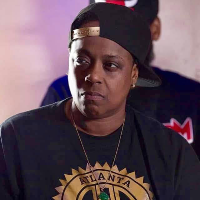Woman wearing a black cap backwards, black shirt with gold logo, and a necklace, looking to the side.