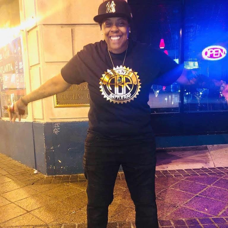 Person smiling with arms outstretched, wearing a black shirt with a gold logo, black pants, and a black cap, standing outside at night.