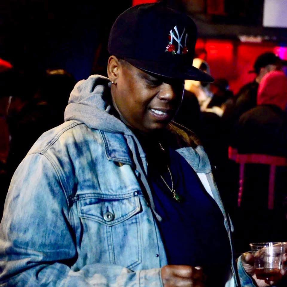 Person wearing a denim jacket and a black cap, smiling in a dimly lit indoor setting with people in the background.