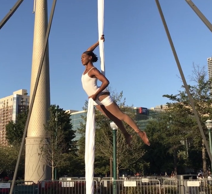 A woman performs aerial silk acrobatics outdoors, hanging horizontally with one arm holding the silk, against a cityscape background.