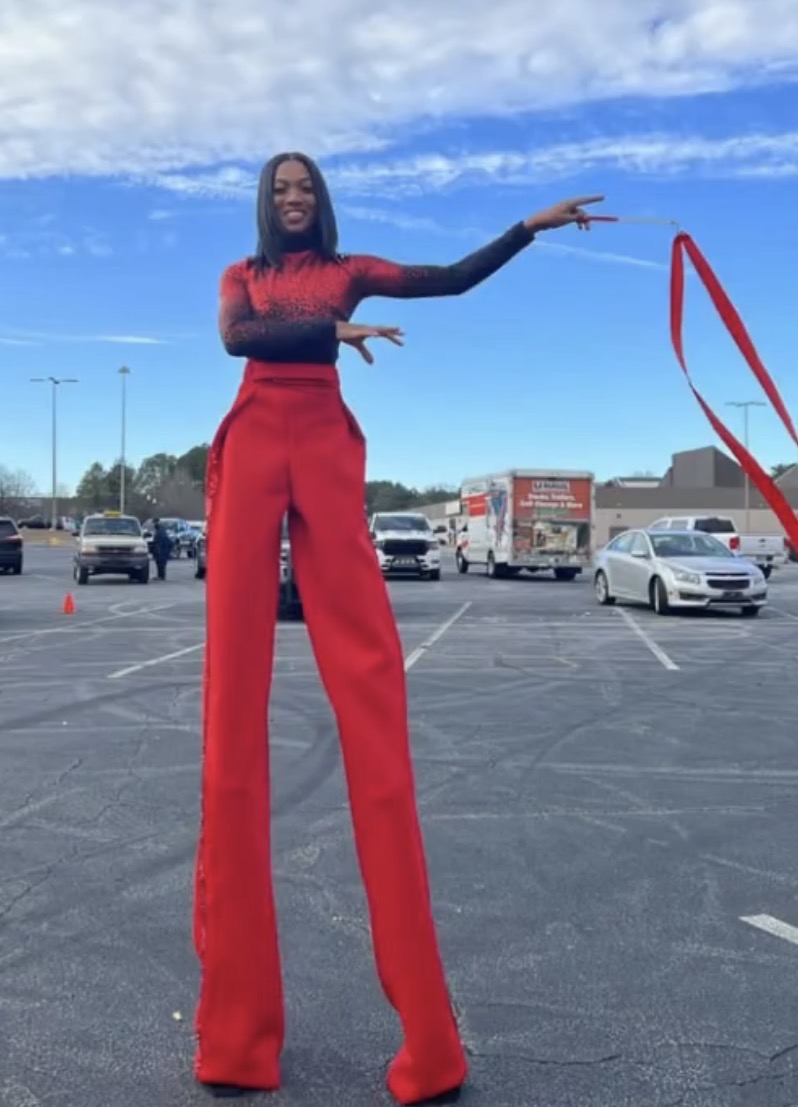 Tall woman in red pants and black top standing in a parking lot with cars and a blue sky background.