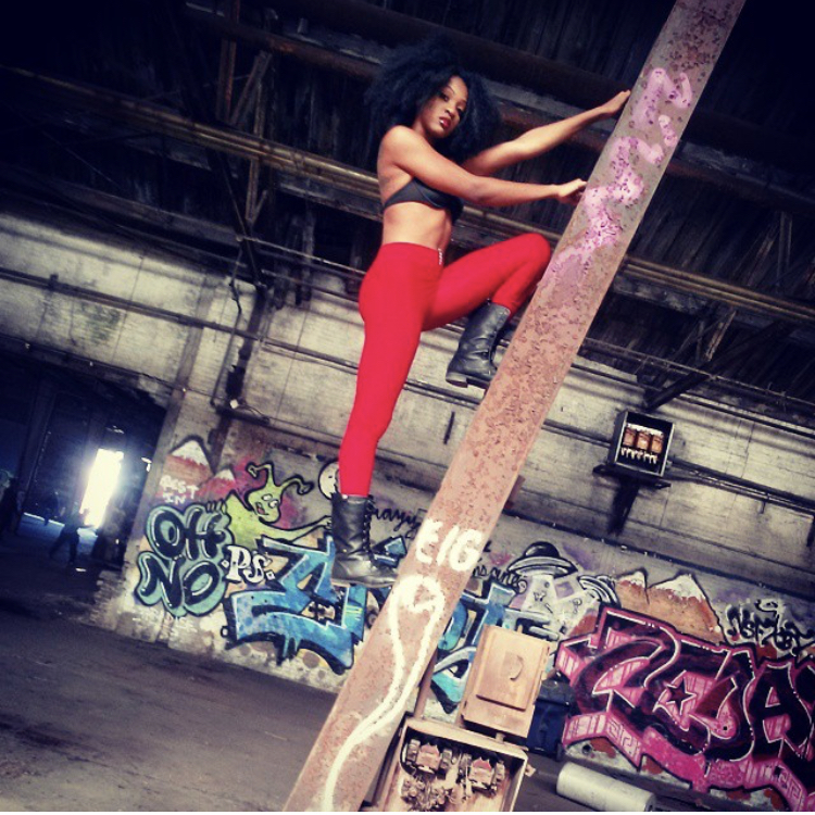 Woman climbing a metal pole in an industrial space with graffiti-covered walls, wearing red pants and black top.