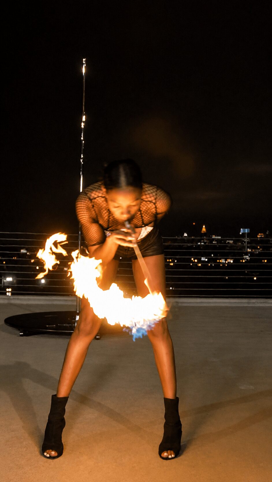 Person performing fire spinning on rooftop at night, with city lights in background.