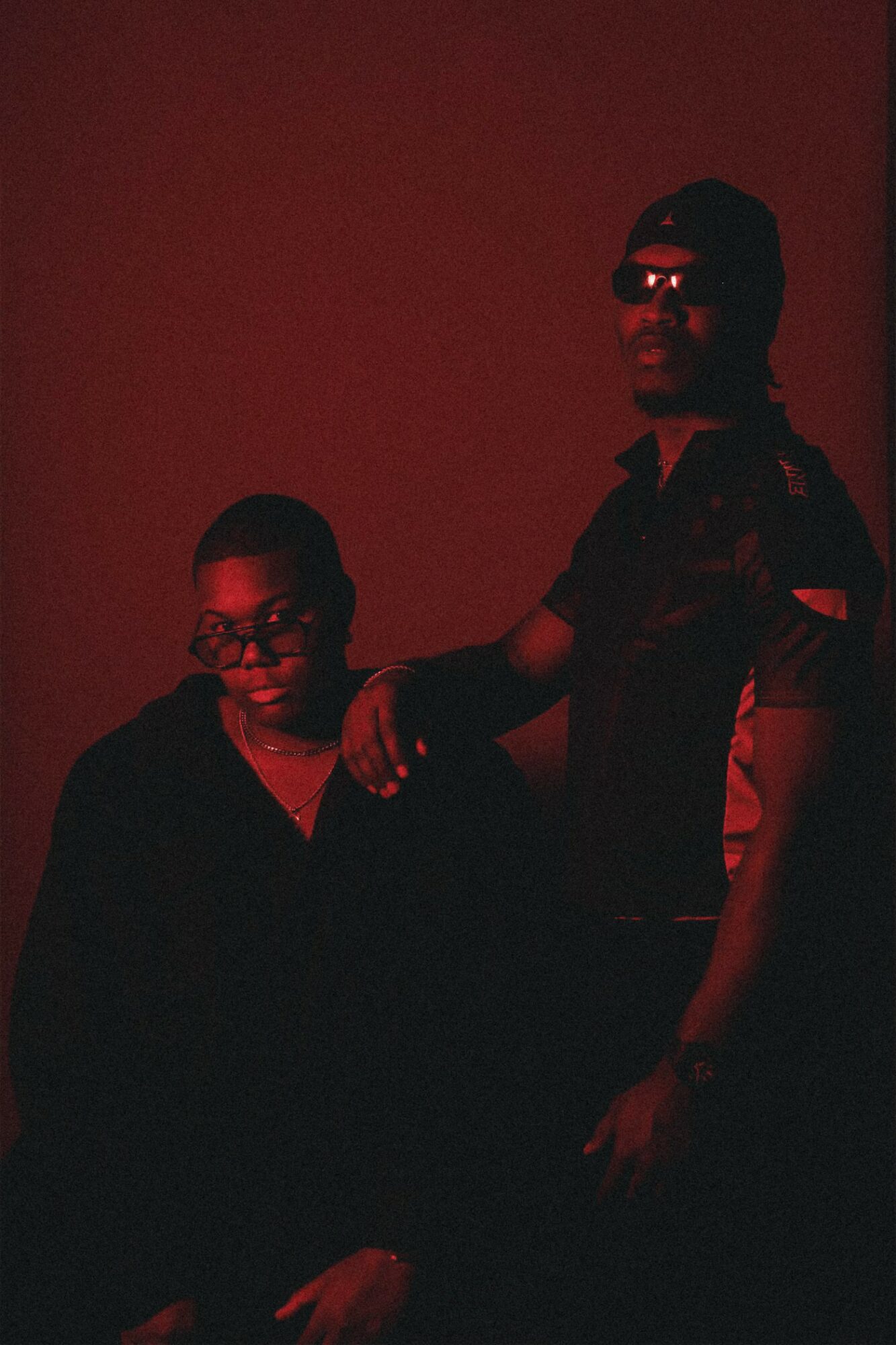 Two people wearing glasses and dark clothing stand against a dark red background.