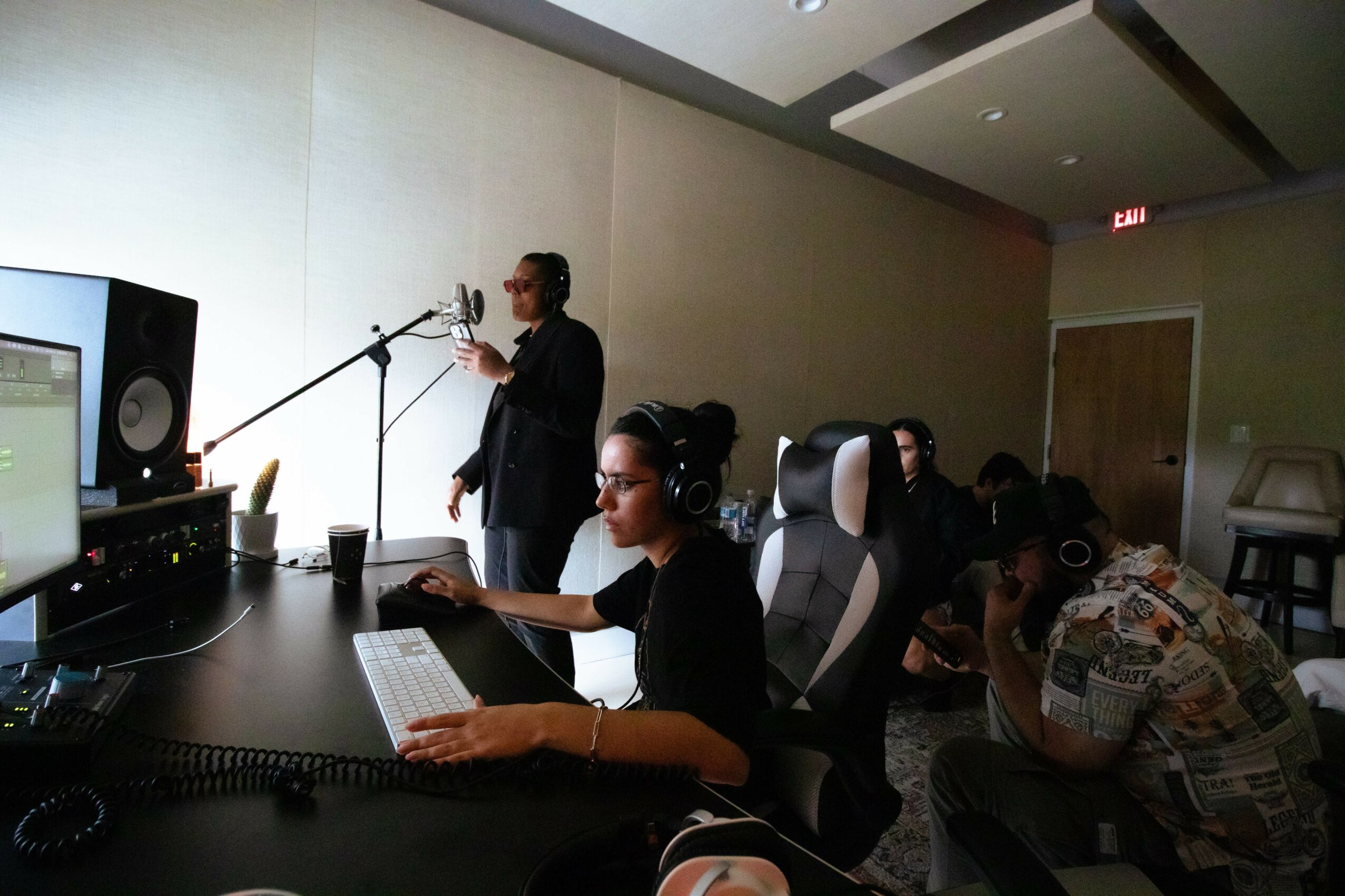 Person in a recording studio working at a computer, with others in the background, some using headphones, in a dimly lit room.