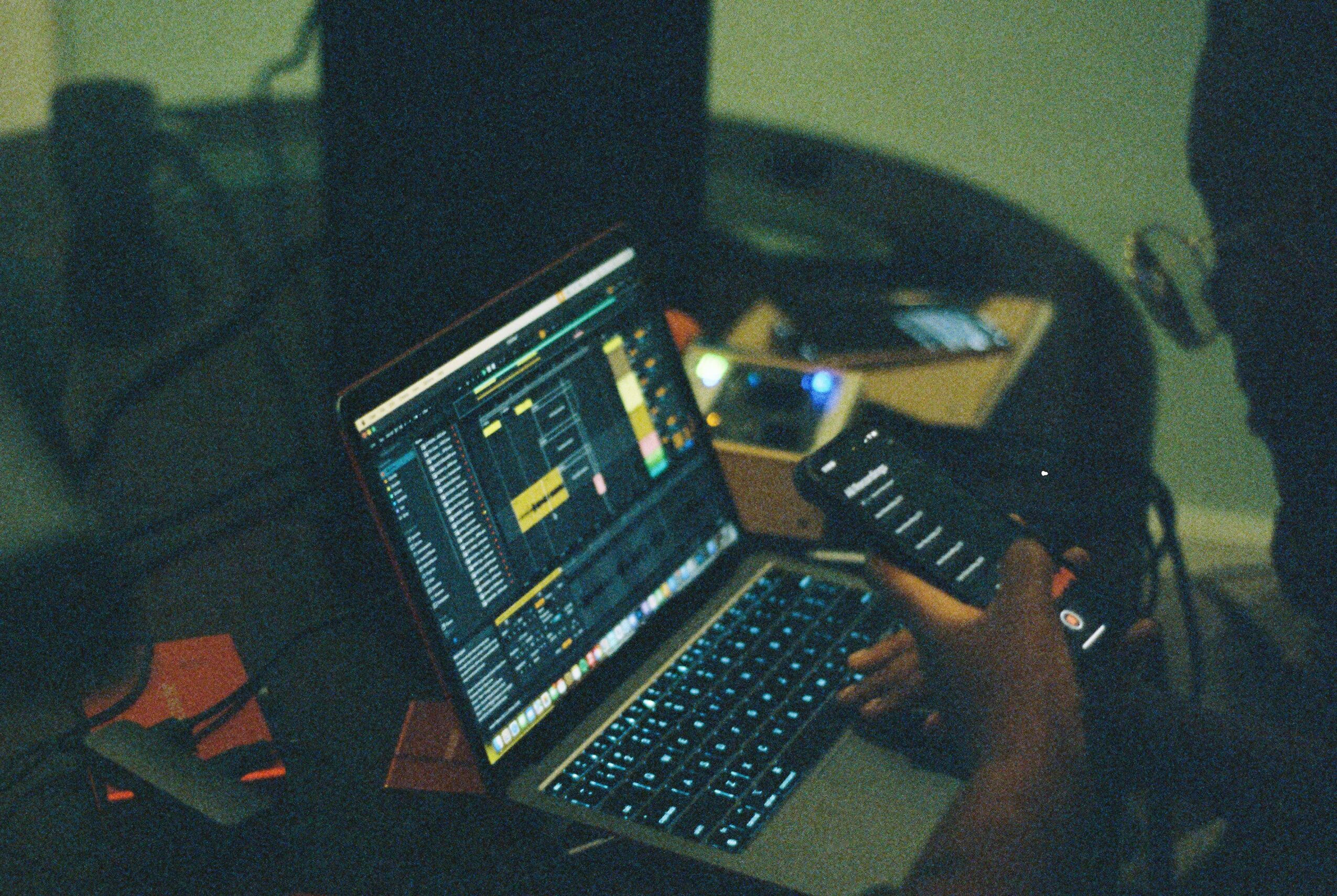Person using a laptop with music production software, holding a smartphone, in a dimly lit room.