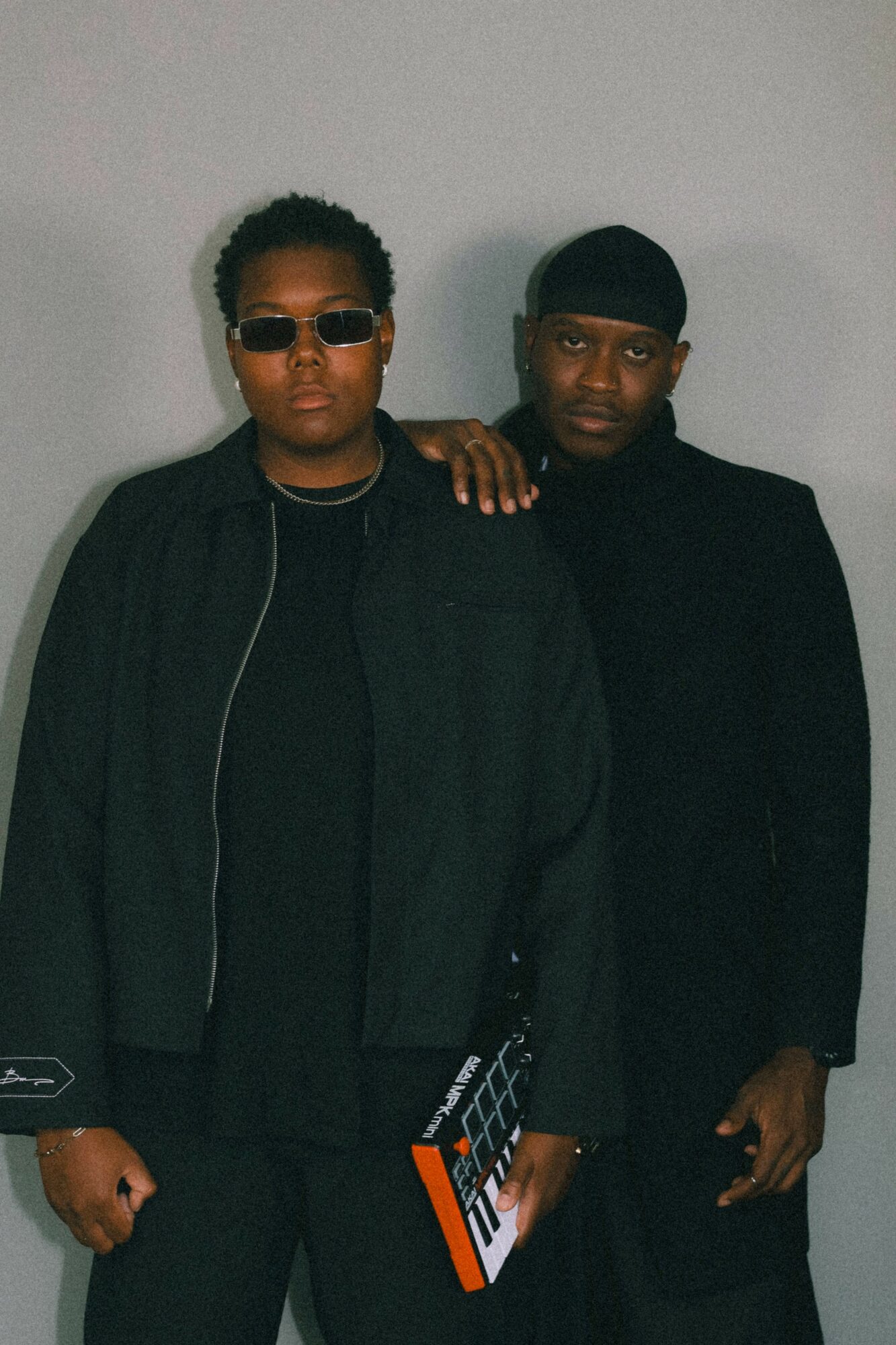 Two people stand side by side, one wearing sunglasses and a black jacket, the other in a black outfit with a head covering.