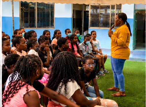 Group of children seated indoors listening to a woman in a yellow jacket speaking, with windows in the background.