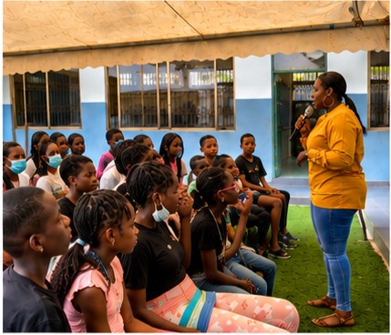 Woman speaking to children seated outdoors, some wearing masks, in a schoolyard with blue walls and a canopy.