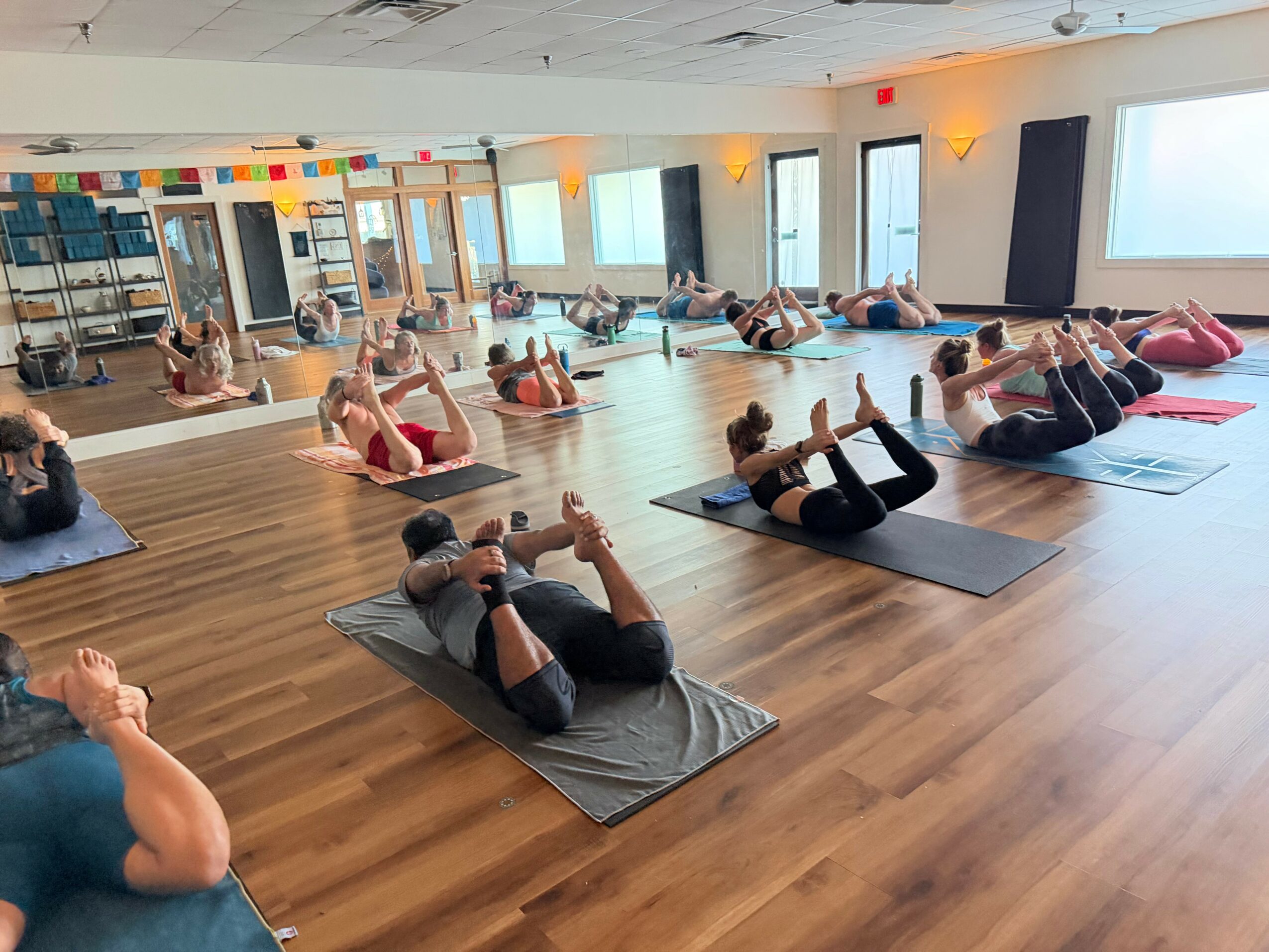 Group of people practicing yoga on mats in a spacious, well-lit studio with large windows and wooden floors.