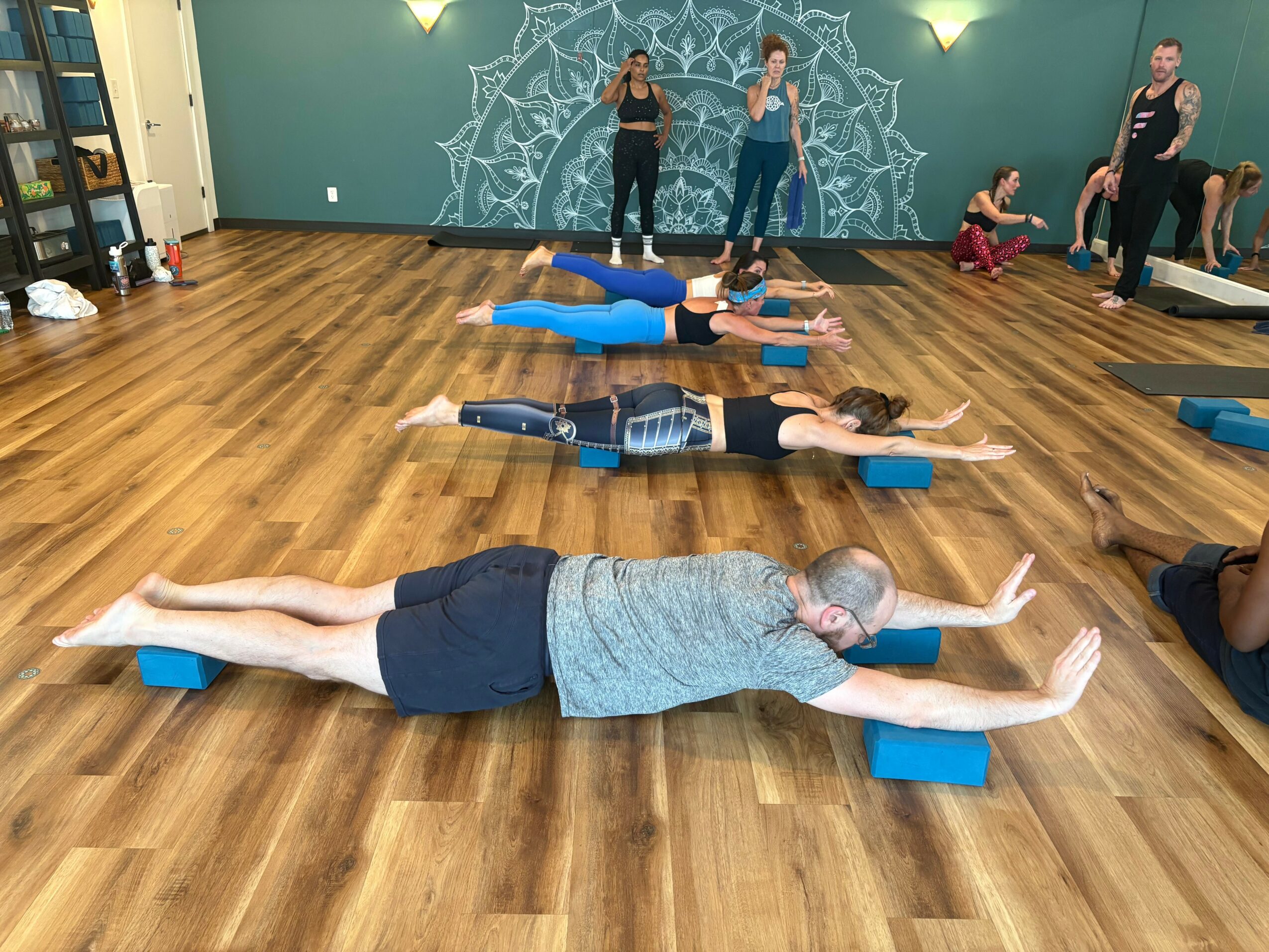 People lying on their stomachs on yoga blocks with arms extended forward in a yoga class, wooden floor, teal wall, and others standing in the background.