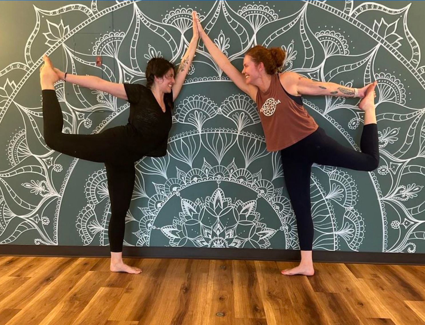 Two women perform a yoga pose with their hands joined above their heads, standing on one leg each, against a patterned wall.