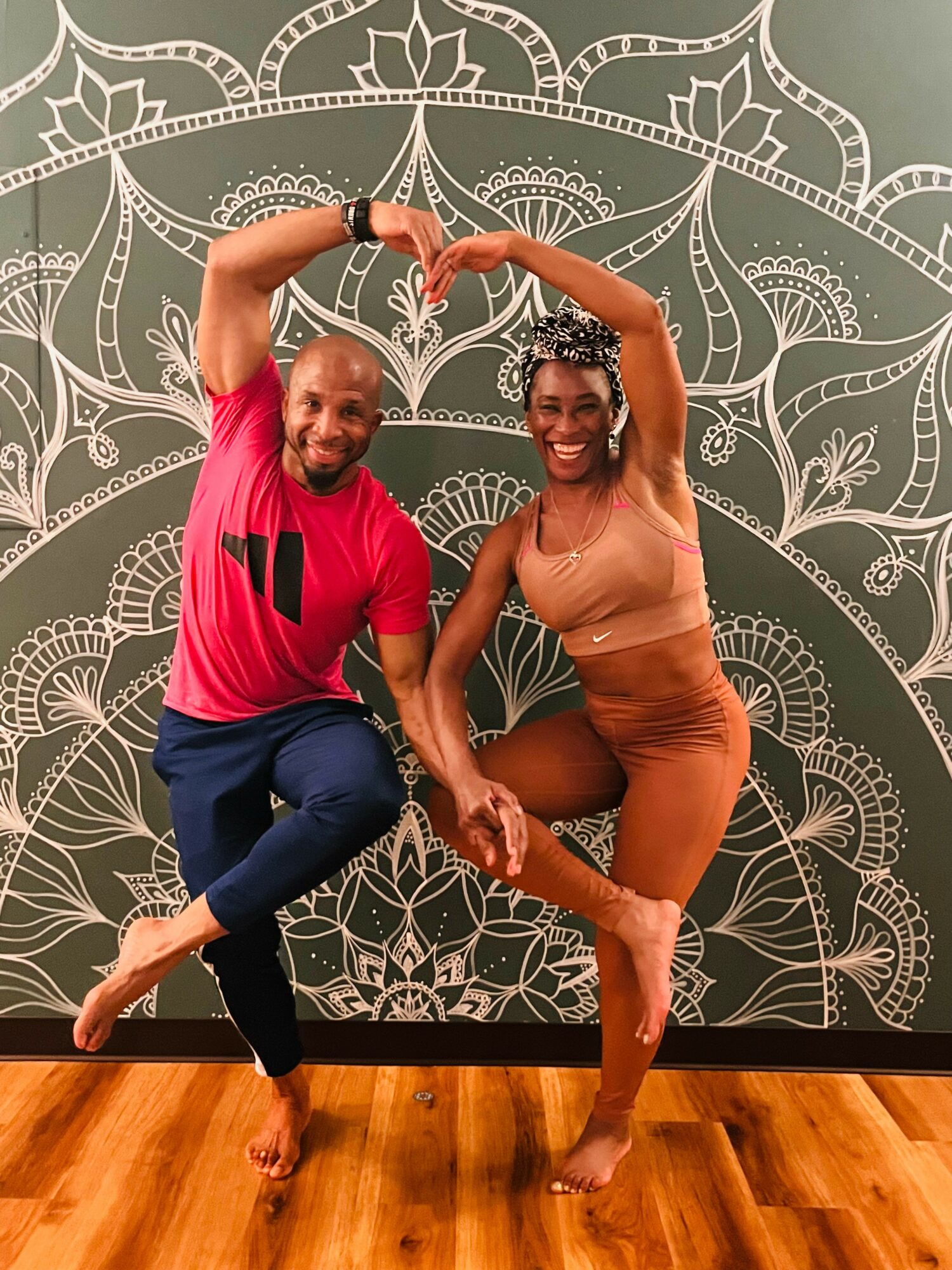 Two people pose with arms forming a heart shape, smiling, in front of a decorative wall pattern, on a wooden floor.