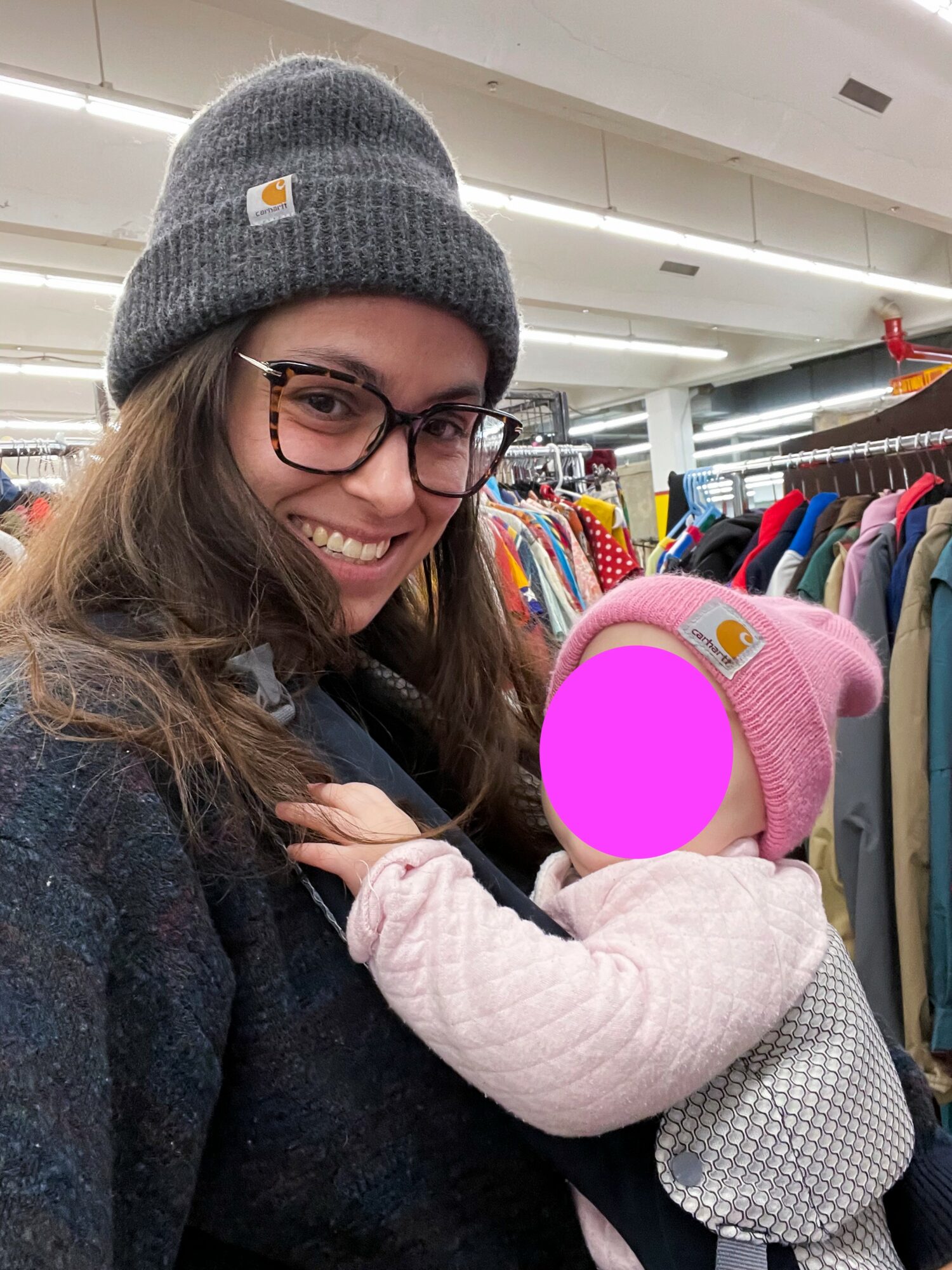 Woman wearing glasses and a gray beanie holding a baby in a pink hat in a clothing store.