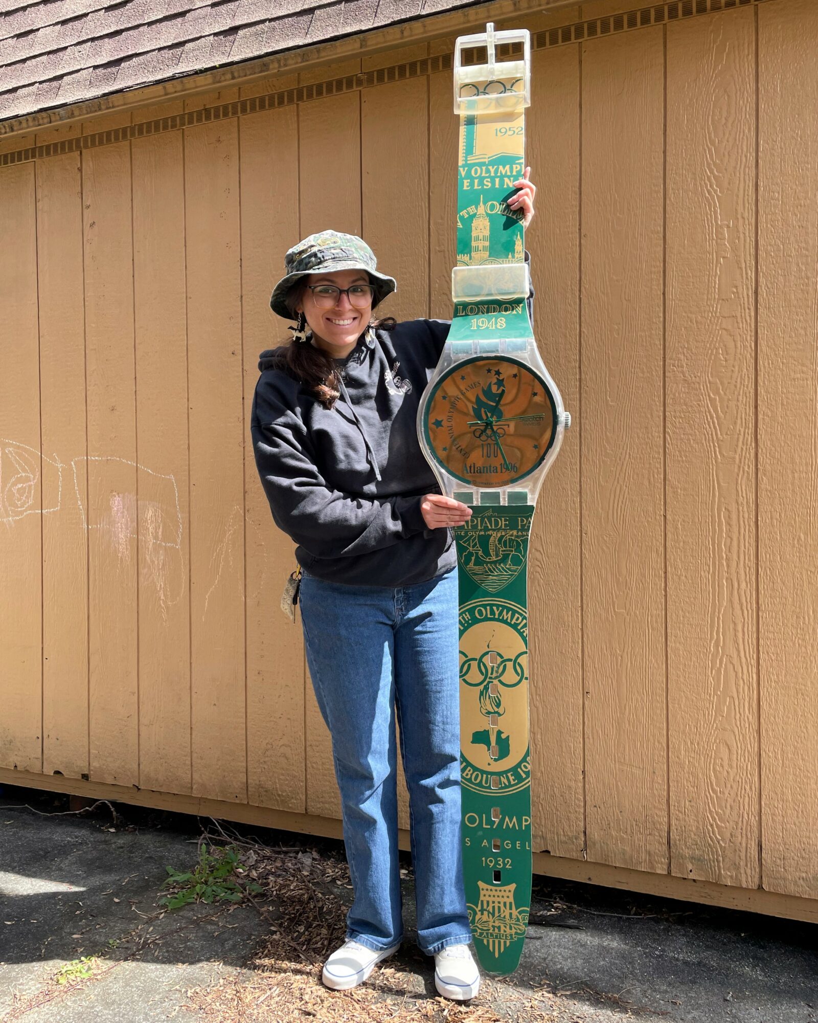 Woman wearing a hat and sunglasses holding a large colorful clock and a tall decorative object outdoors, standing in front of a wooden wall.