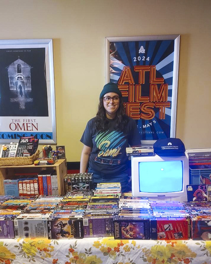 Person standing behind a table with comic books and a vintage computer, smiling, in a room with posters on the wall.