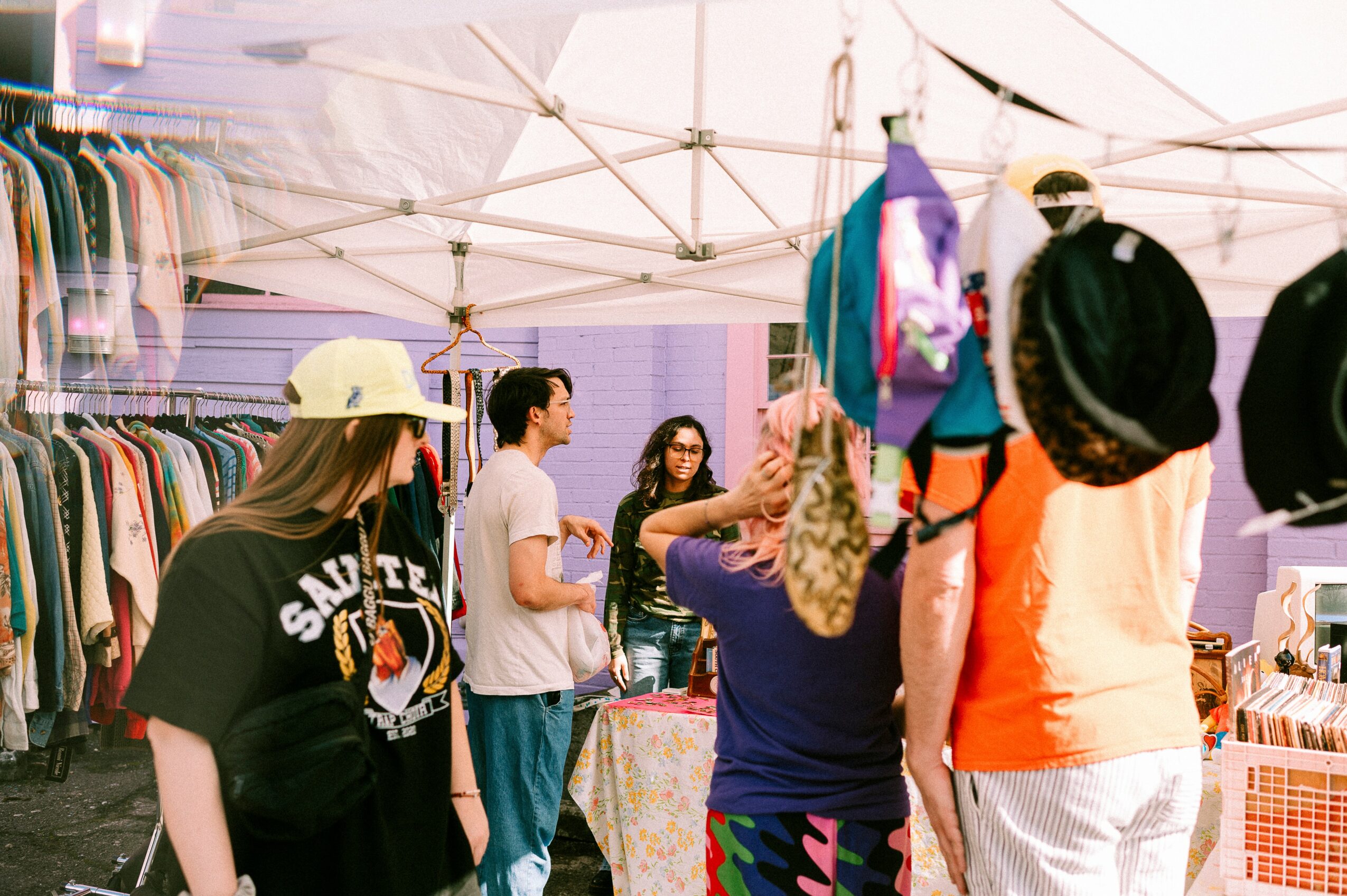 People browsing clothing at an outdoor market stall under a white canopy, with racks of clothes and accessories visible.